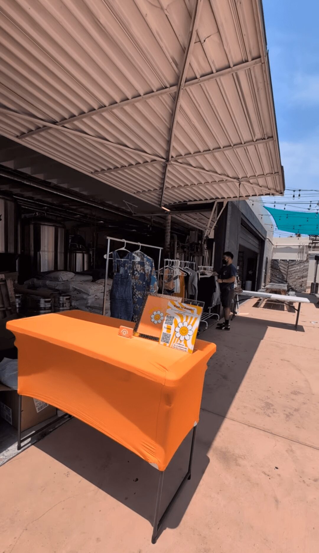 Orange table with a colorful sign, set outside under a large beige canopy, with buildings and a blue sky in background.