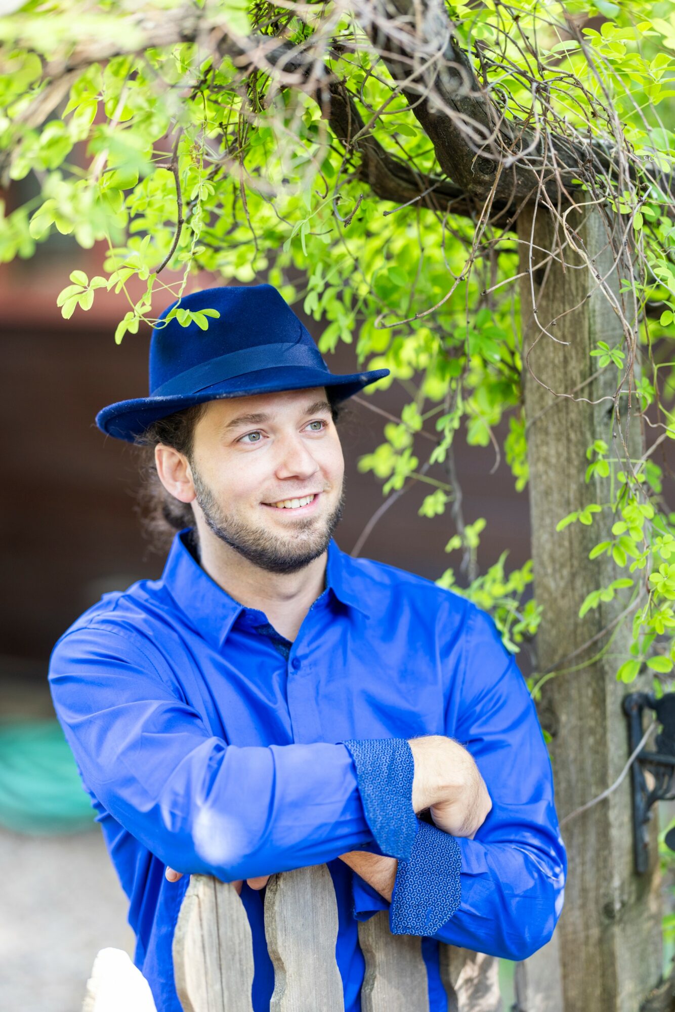 Young man with a beard wearing a blue shirt and hat, standing outdoors near a tree with green leaves.