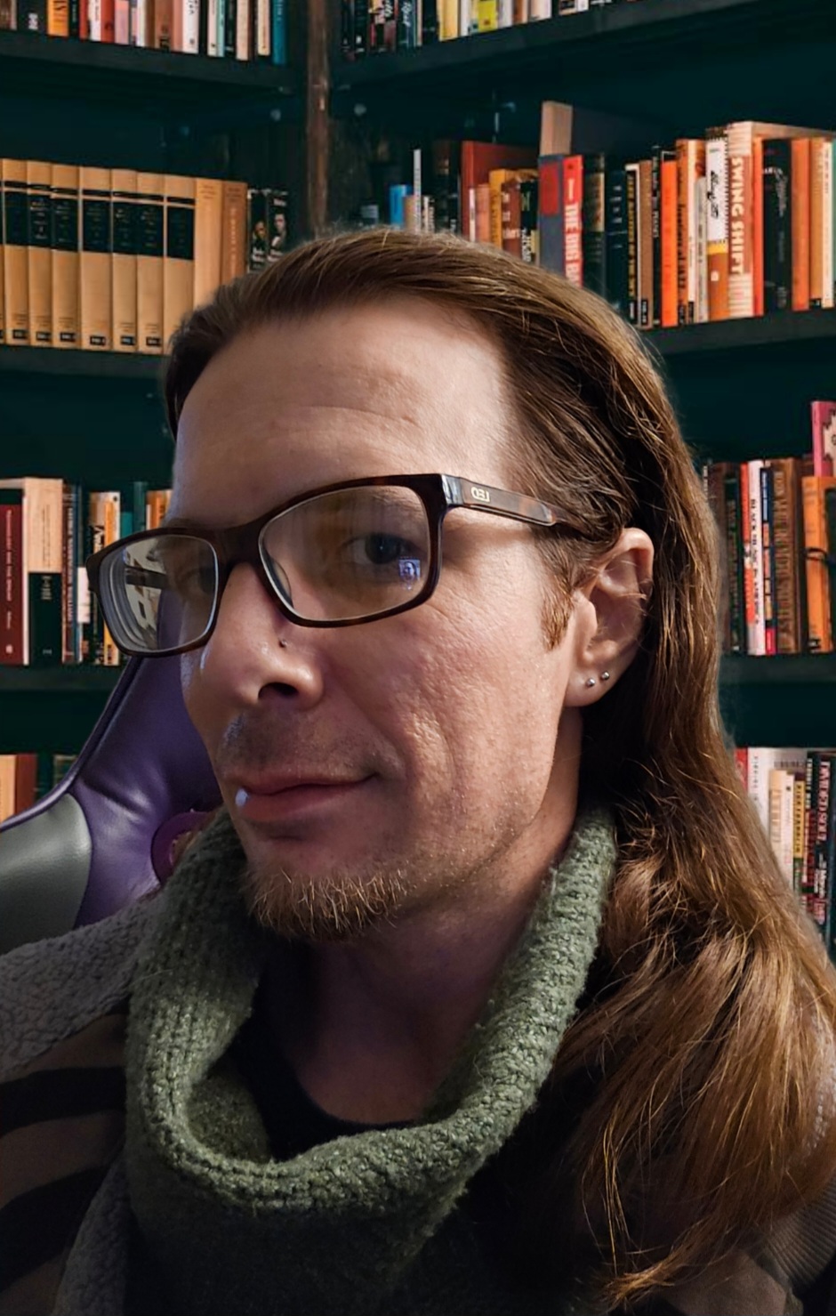 Person with glasses and long hair in a library, looking at the camera, bookshelves in the background.