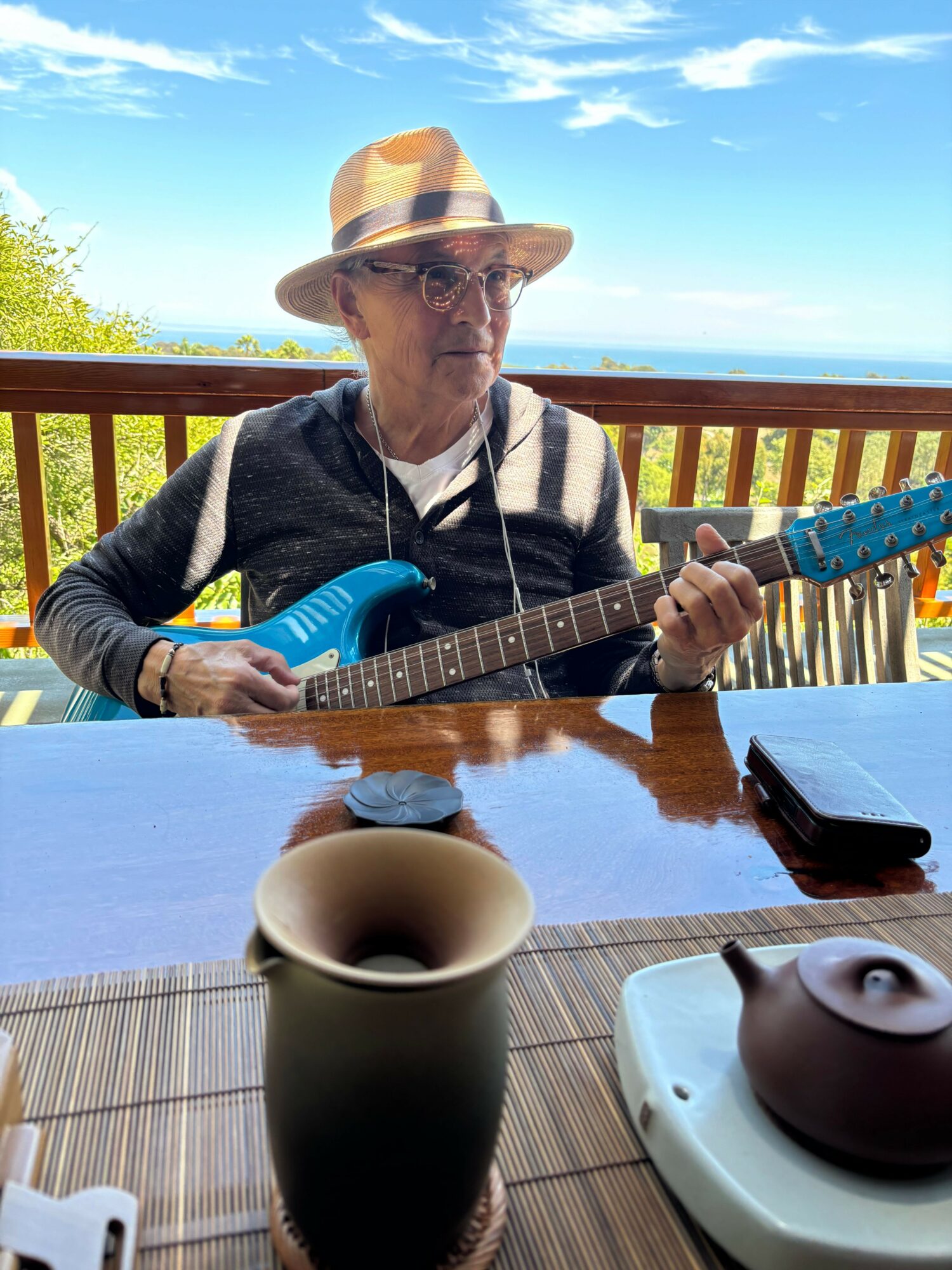 Person wearing a hat playing guitar at outdoor table with a cup and speaker, sunny sky background.