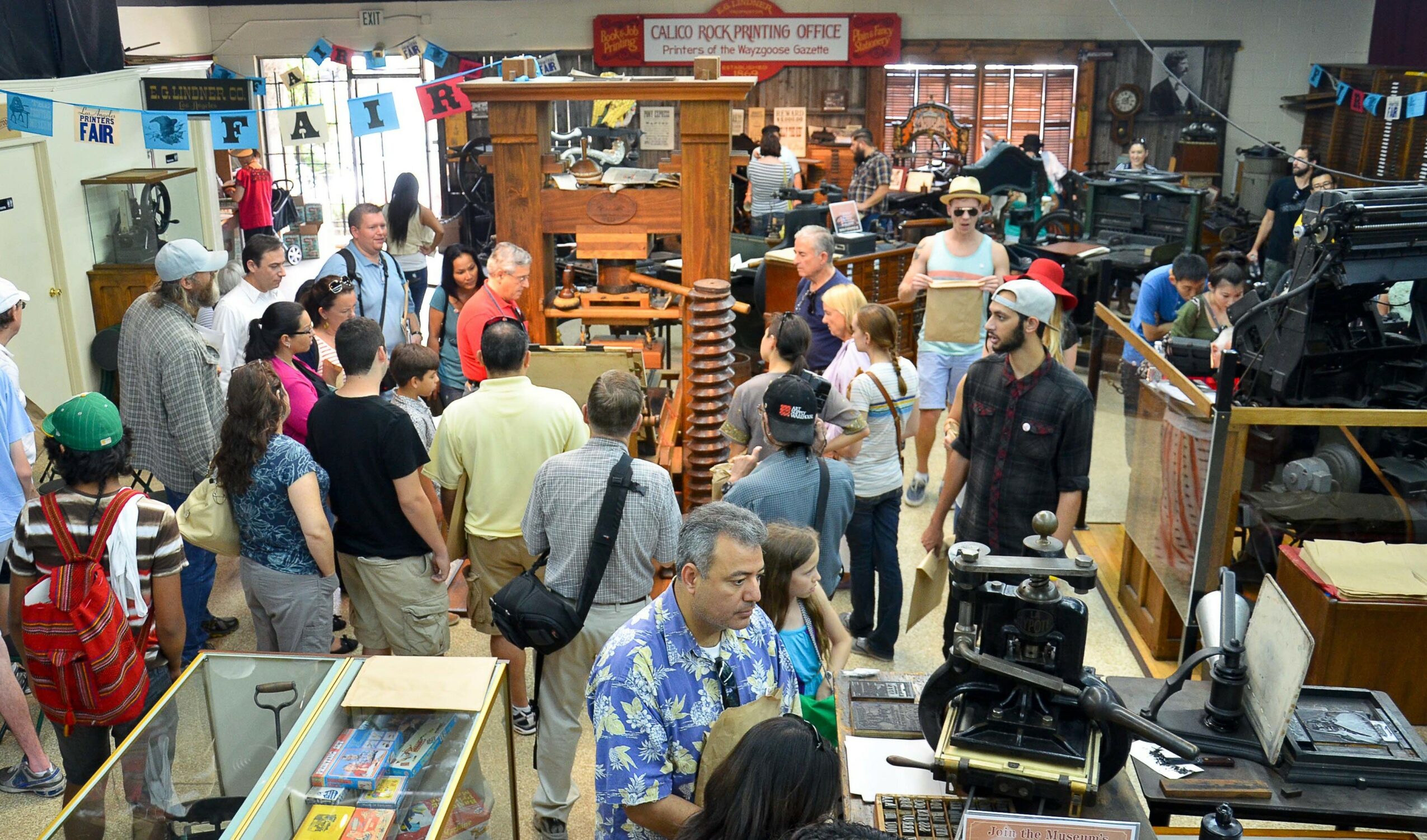 Crowd of people browsing books and items in a bookstore or library, with shelves and displays visible.