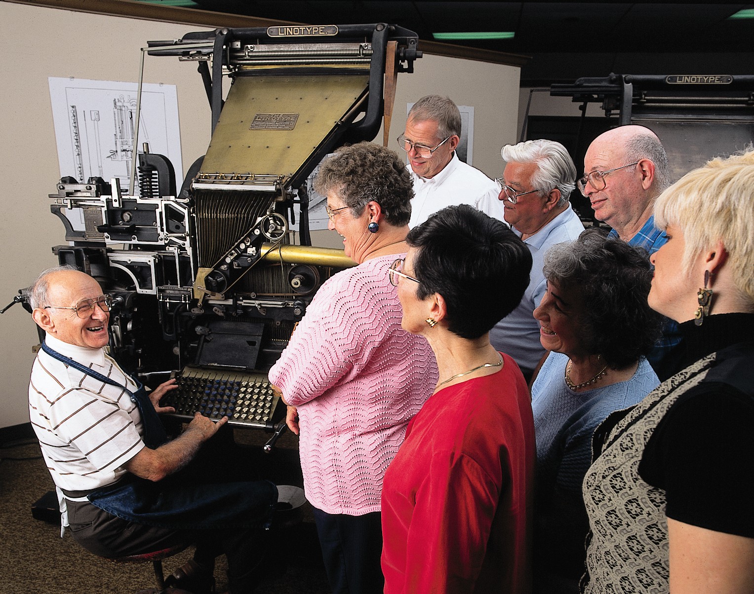 Group of people gathered around a person operating a large printing machine, smiling and engaging with each other.