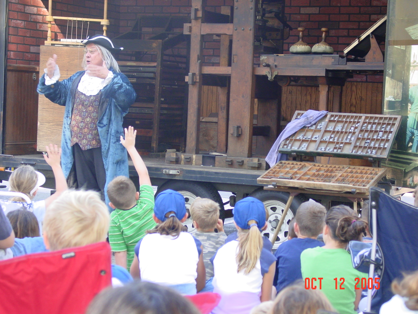 Person dressed as a historical figure speaking to children outdoors, with children raising hands, on a stage with props.