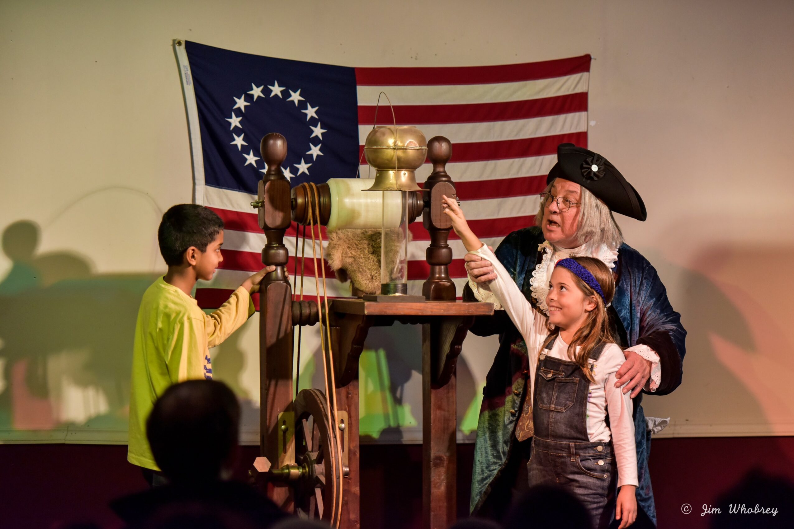 A woman dressed as a historical figure demonstrates a spinning wheel to two children in front of an American flag backdrop.