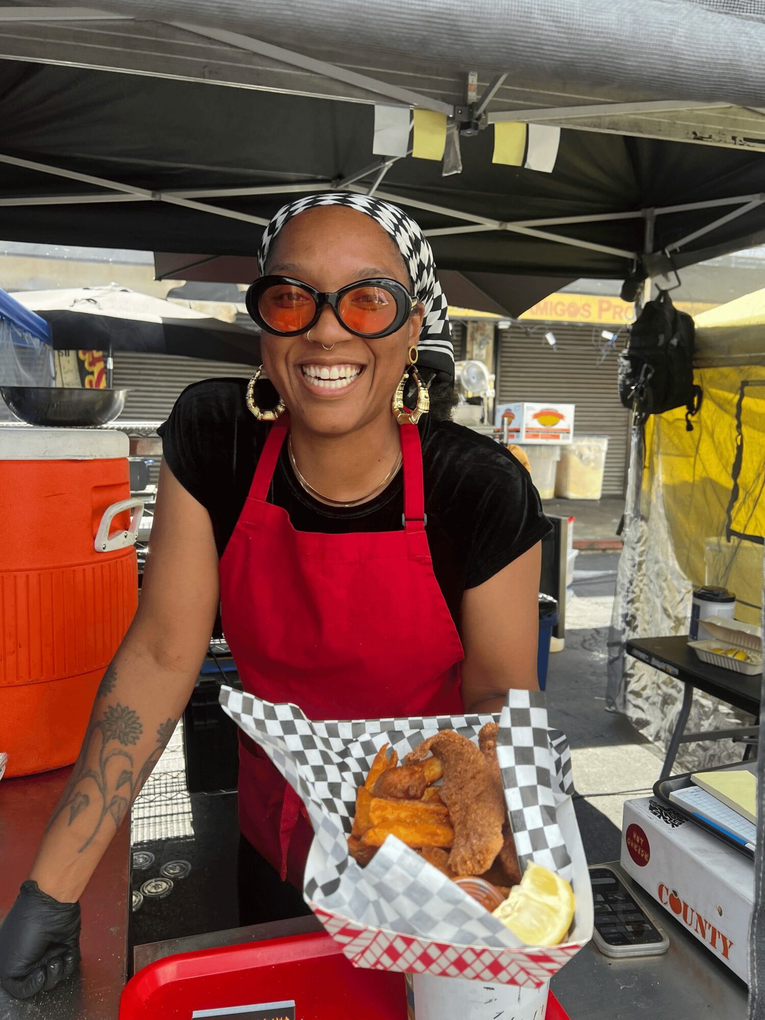 Smiling woman wearing sunglasses, a checkered headscarf, red apron, and black shirt, standing at a food stall with fried food and lemon slices.