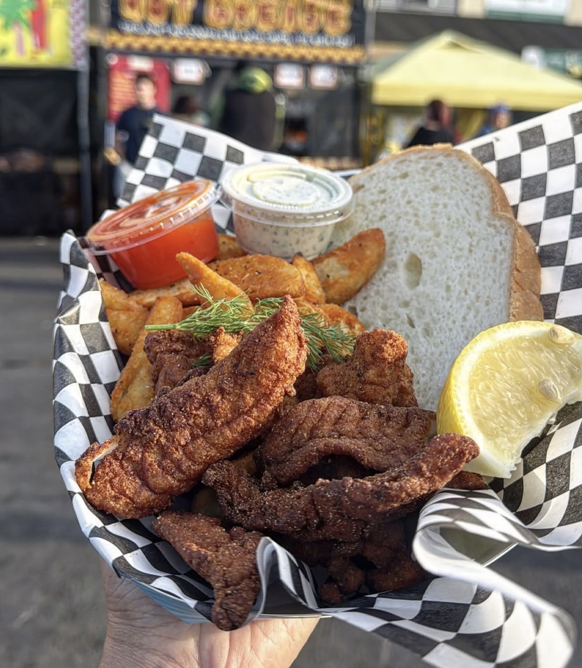 Basket of fried chicken, potato wedges, bread, lemon wedge, and dipping sauces at an outdoor event.