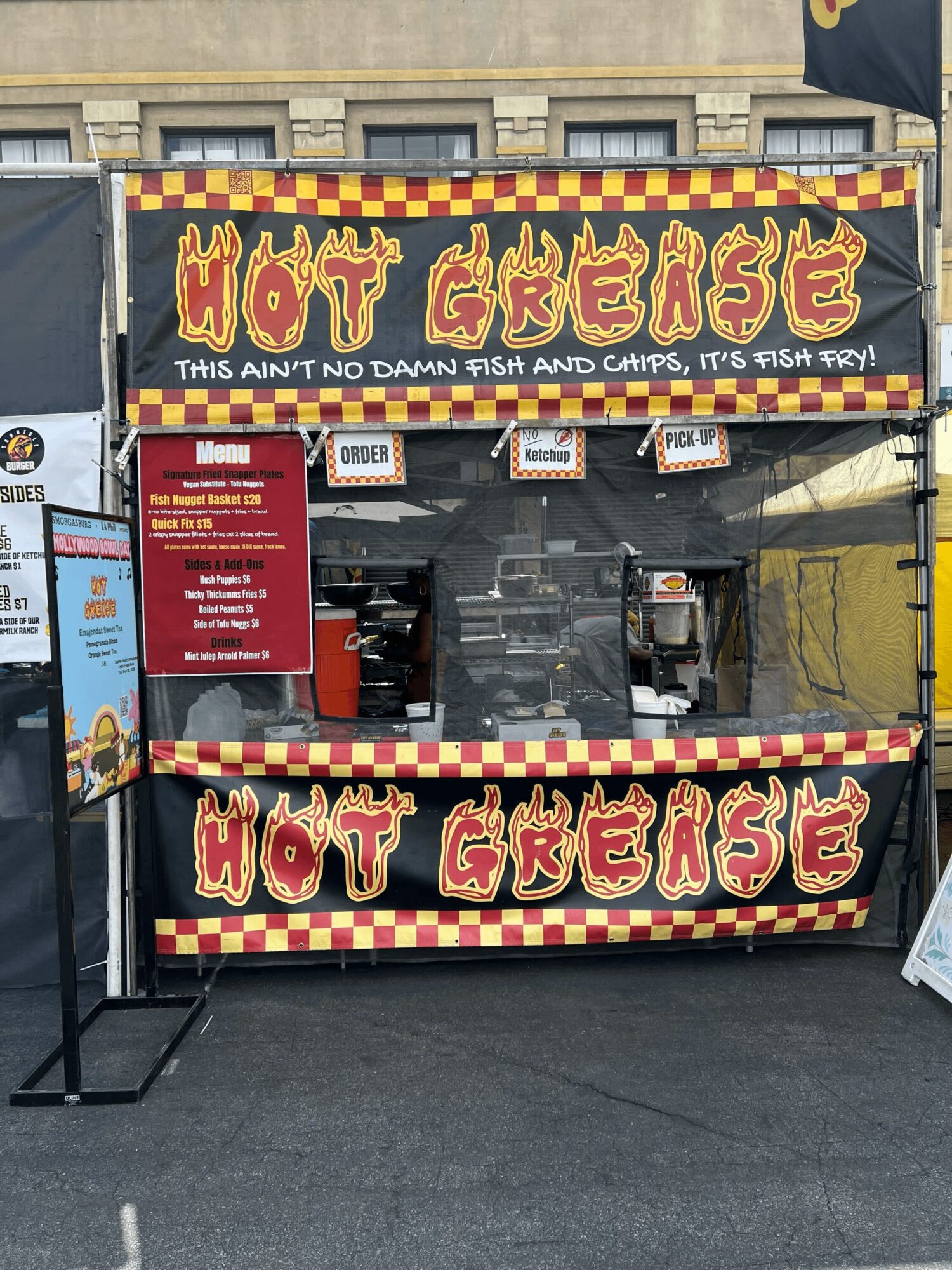 Food stand with signs reading 'Hot Gheease' and a tagline about fish and chips, with a red menu board and black and yellow checkered banners.