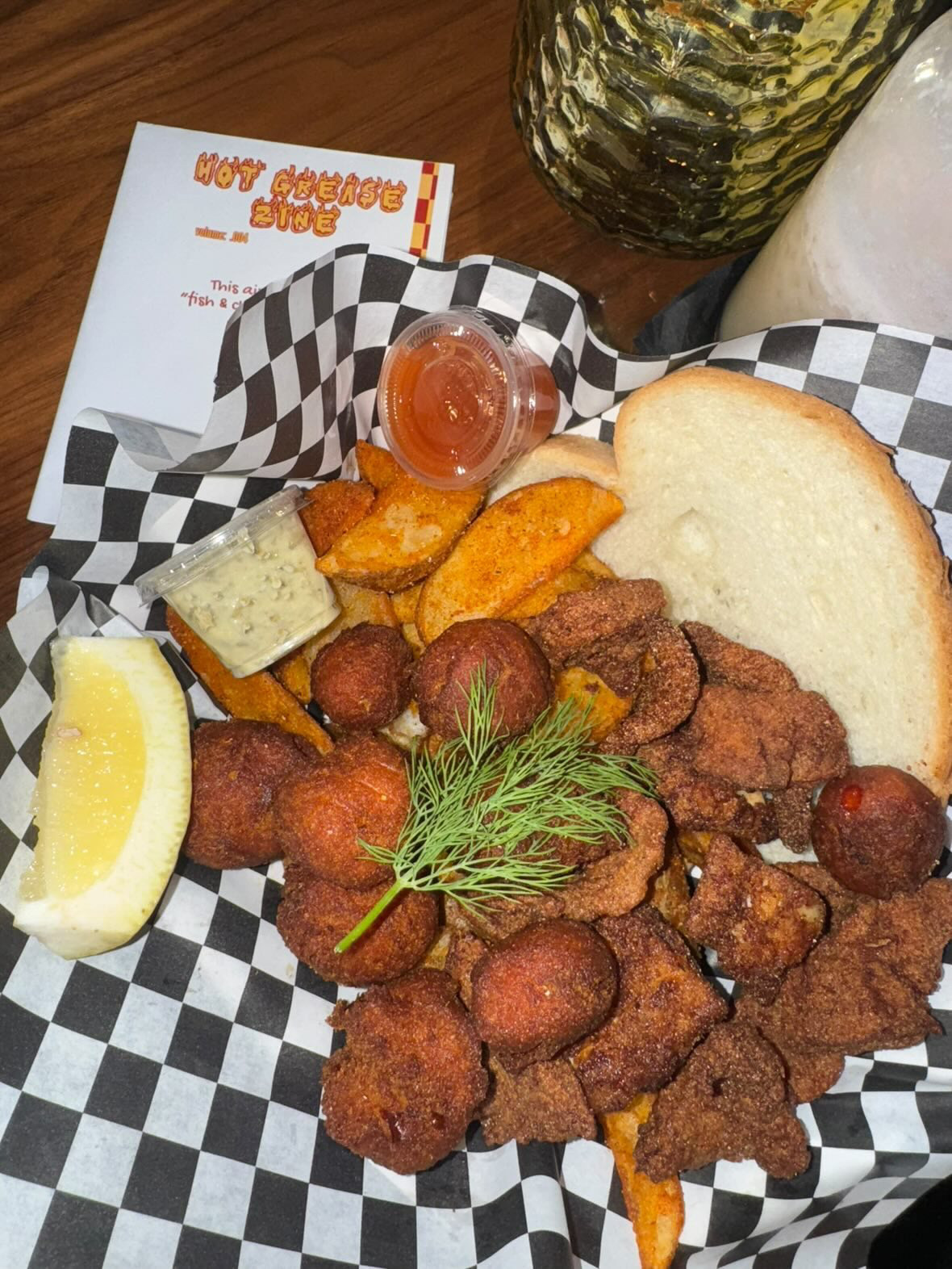 Fried chicken pieces with a lemon wedge, potato wedges, a small cup of sauce, bread, and a sprig of dill on checkered paper.
