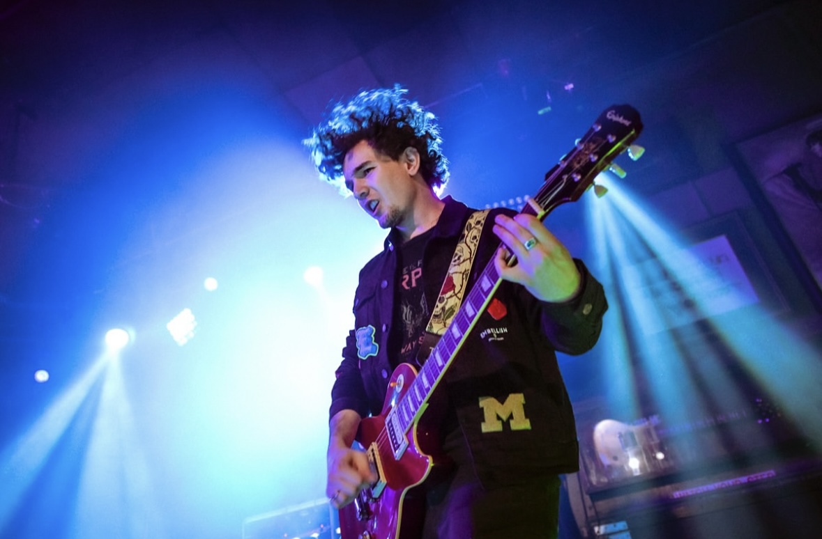 Person playing an electric guitar on stage with colorful lighting effects, curly hair, and a dark jacket.