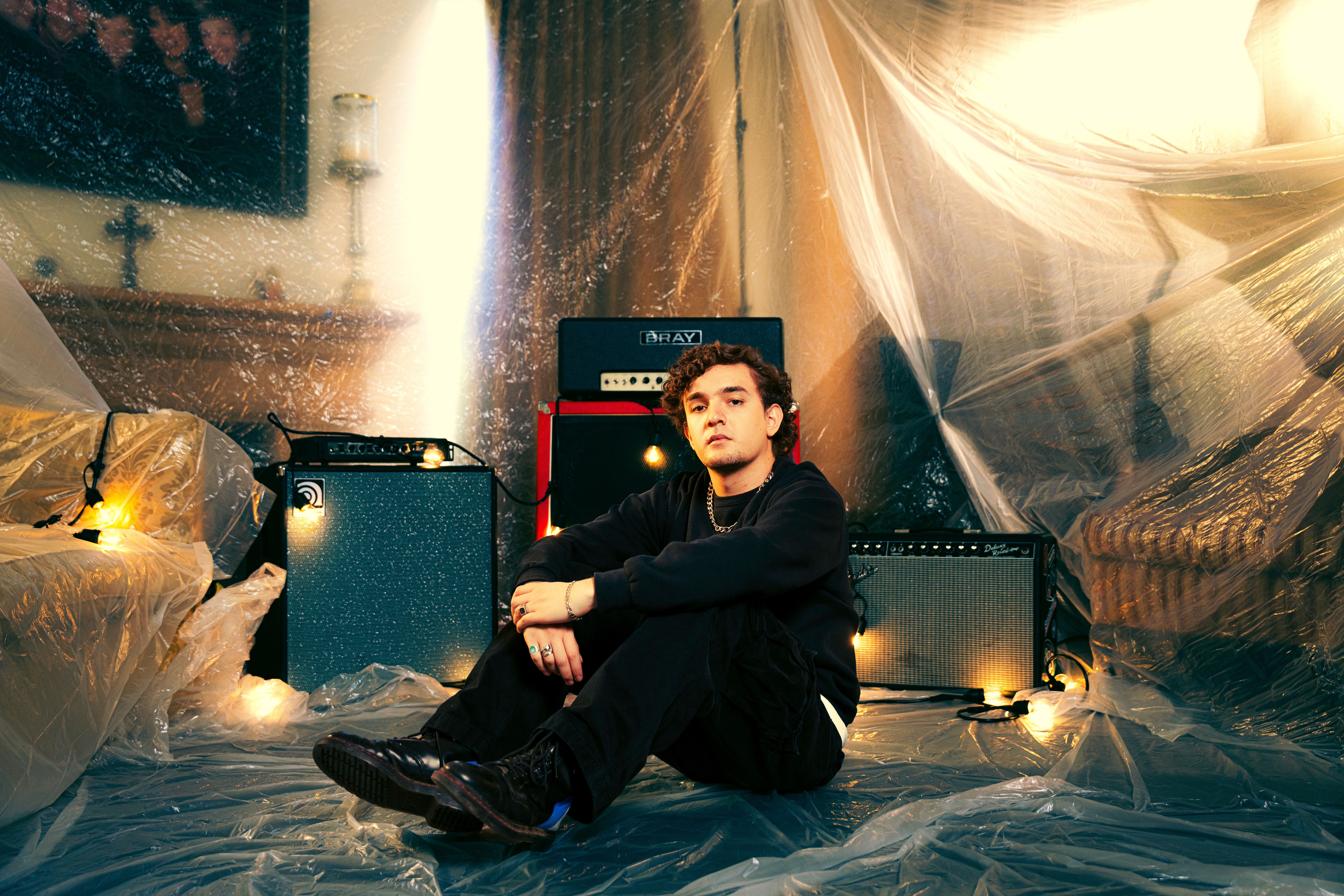 Person sitting on the floor surrounded by musical equipment and plastic-covered furniture in a dimly lit room.