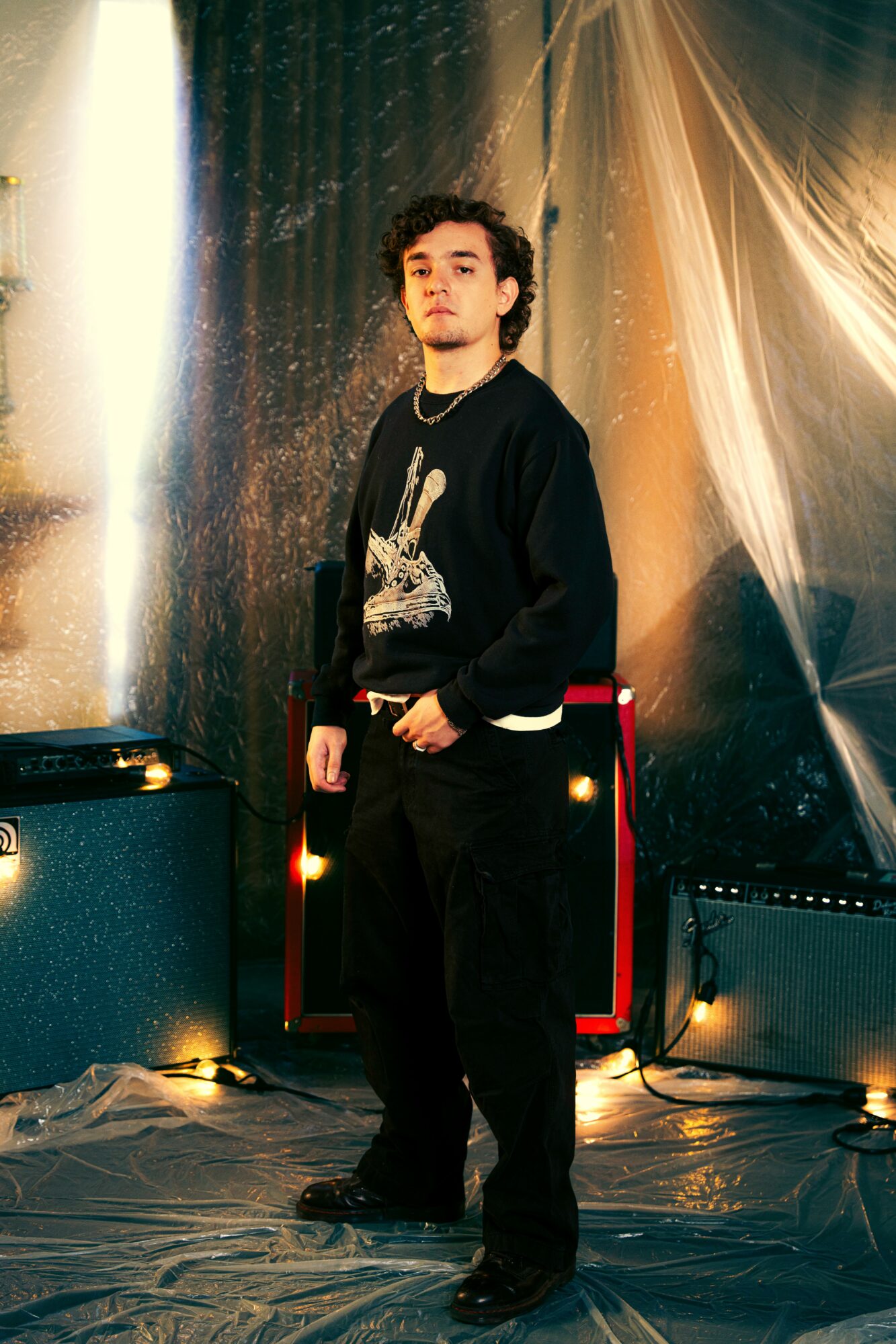 Young man with curly hair stands in front of a shiny backdrop, wearing a black sweater and dark pants.