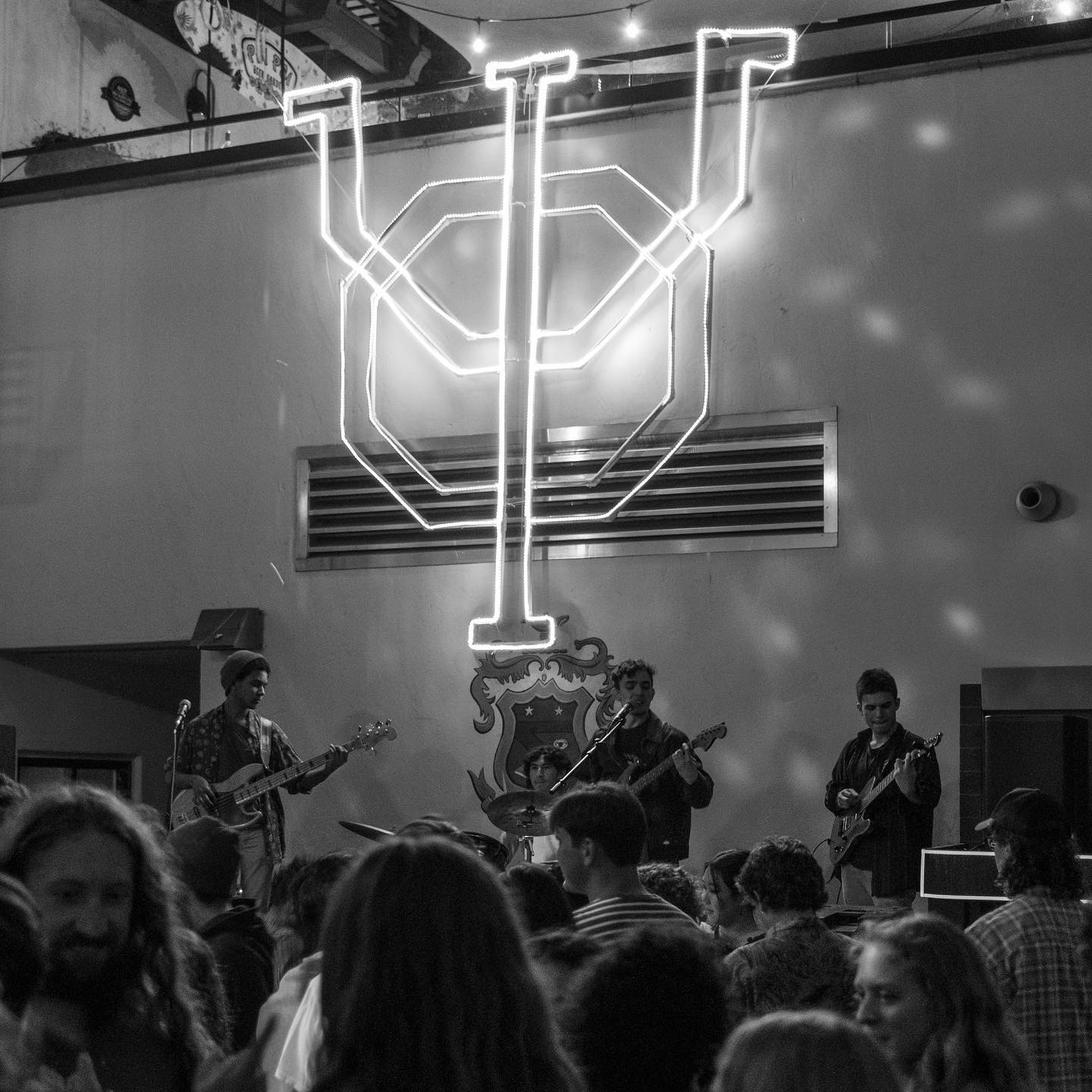 Band performs on stage with audience in front, neon sign above, and a crest on the wall behind.