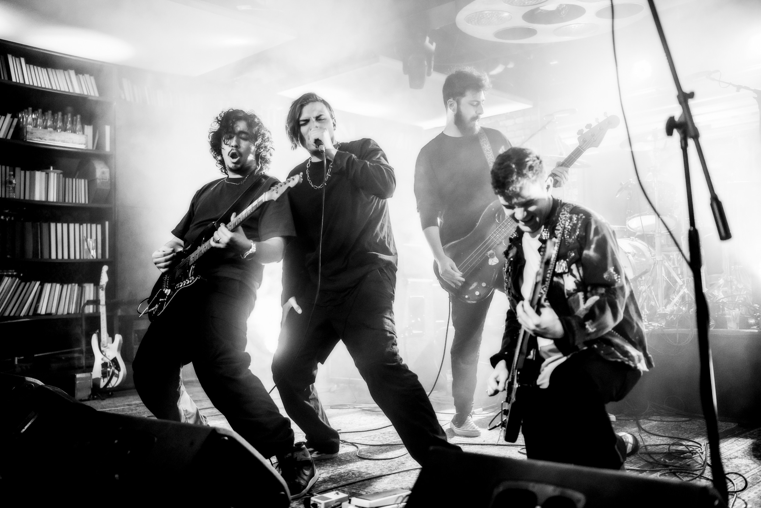 Four musicians perform on stage with guitars and microphone, surrounded by stage lights and equipment.