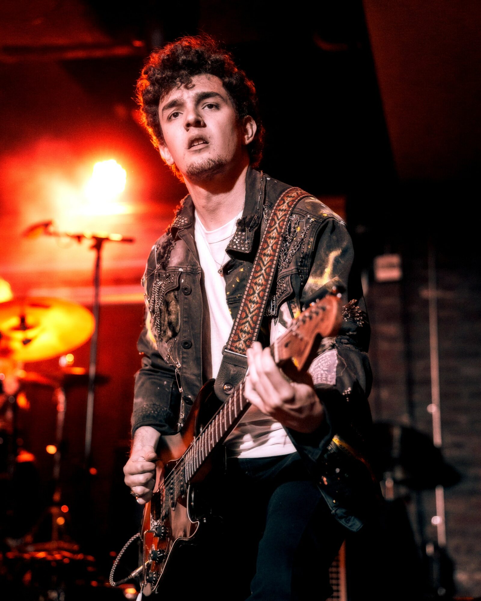 Young man playing electric guitar on stage with red lighting, wearing a denim jacket and white shirt.