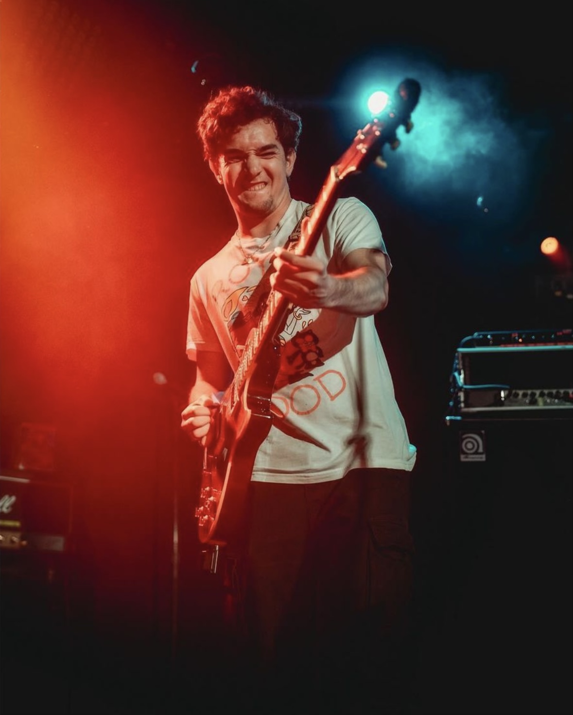 Young man playing an electric bass guitar on stage, smiling, with colorful stage lighting in the background.