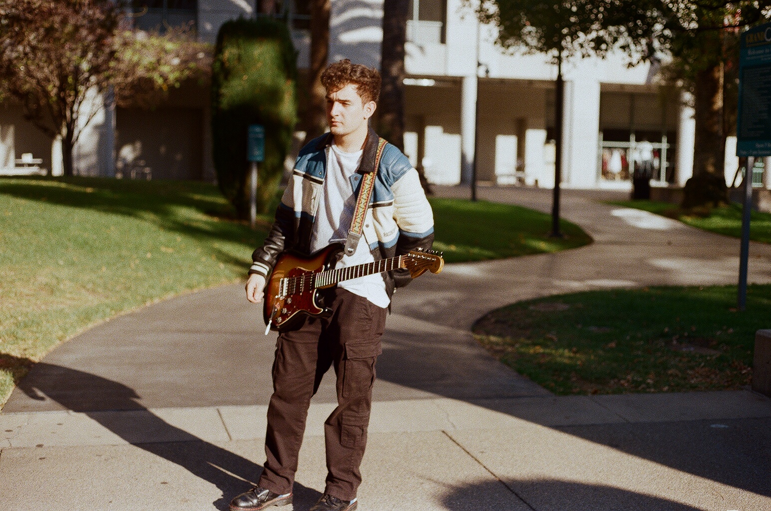 Young person standing on sidewalk holding a guitar, with trees and buildings in the background.