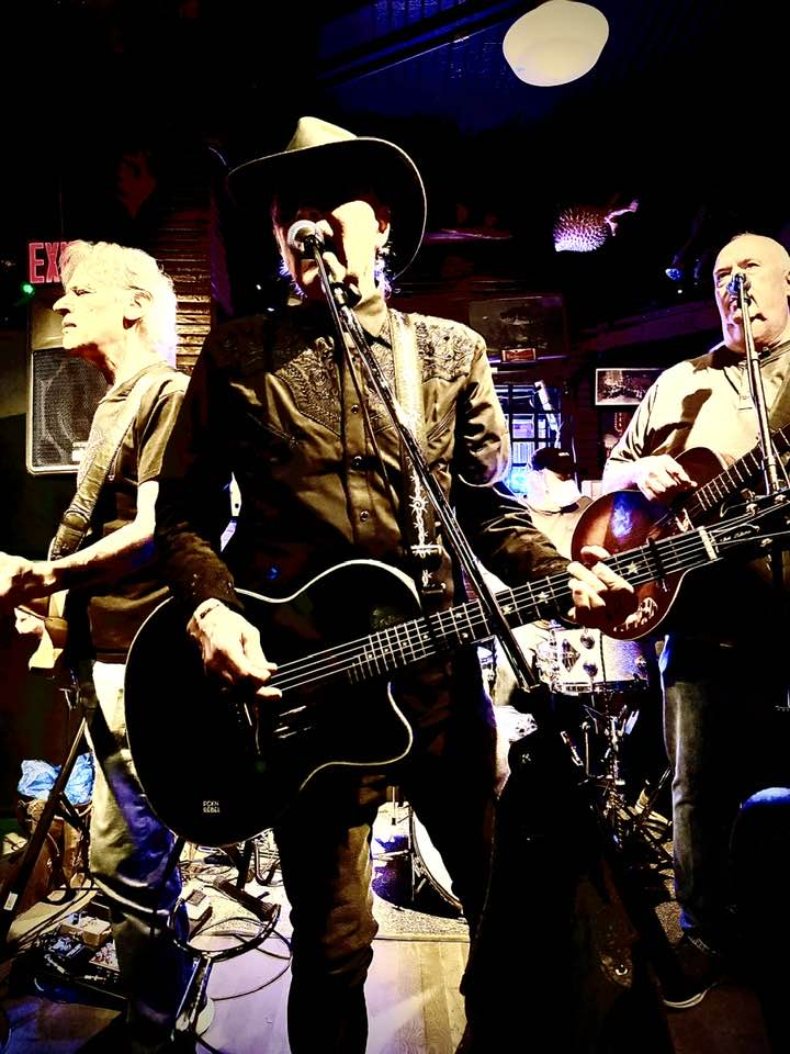 Three musicians perform on stage with guitars, one wearing a hat, in a dimly lit venue.
