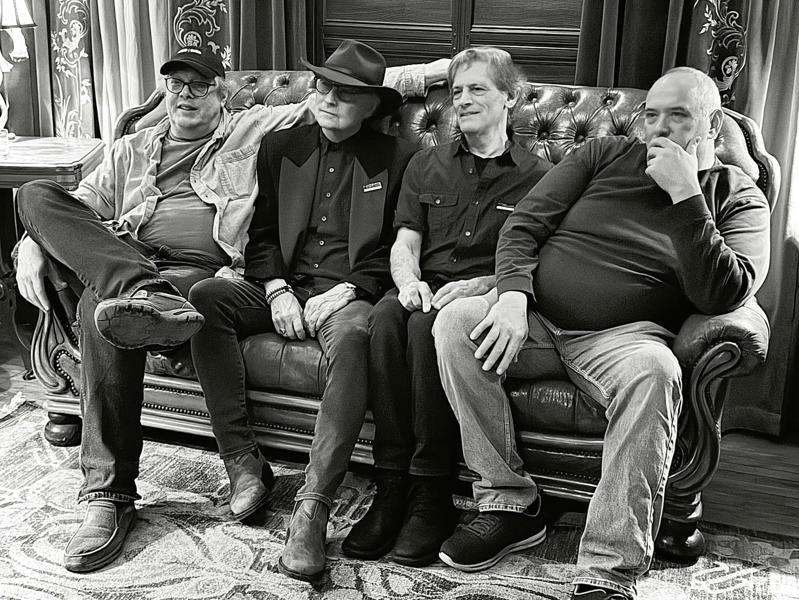 Four men sit on a vintage sofa in a room with wood-paneled walls and curtains, engaging in conversation.