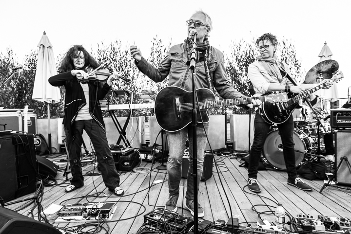 Four musicians perform on an outdoor stage with instruments and equipment, with trees and umbrellas in the background.