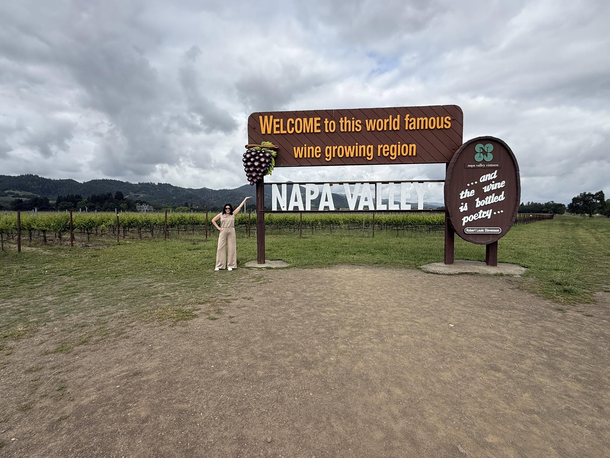 Person standing next to a large welcome sign for Napa Valley wine region, with mountains and cloudy sky in background.