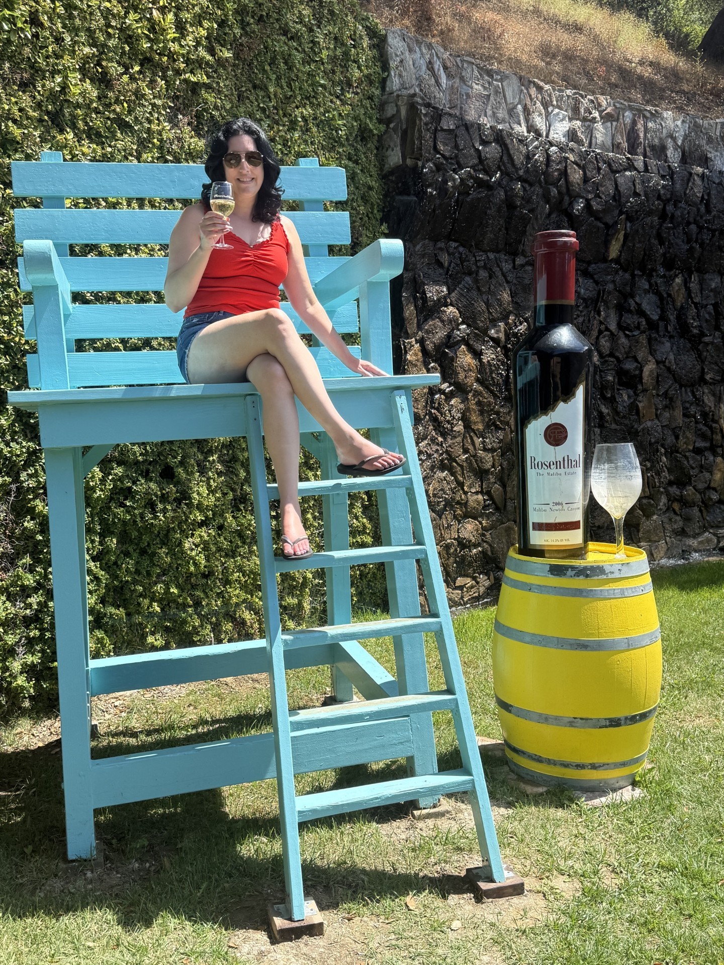 Woman sitting on a large blue chair with a ladder, holding a drink, outdoors with greenery and a stone wall.