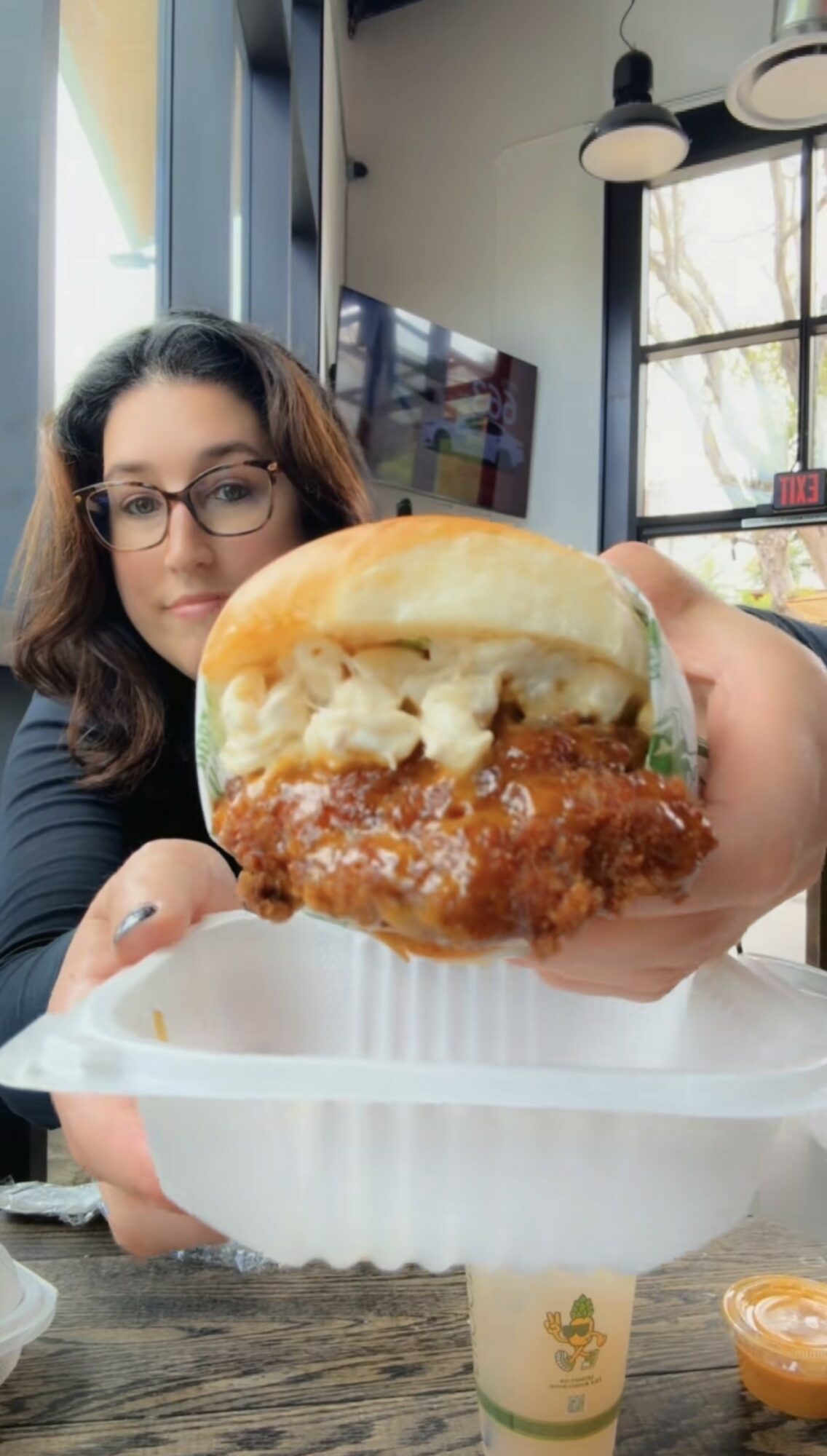 Person holding a sandwich with fried chicken, lettuce, and bun, inside a restaurant with large windows and hanging lights.