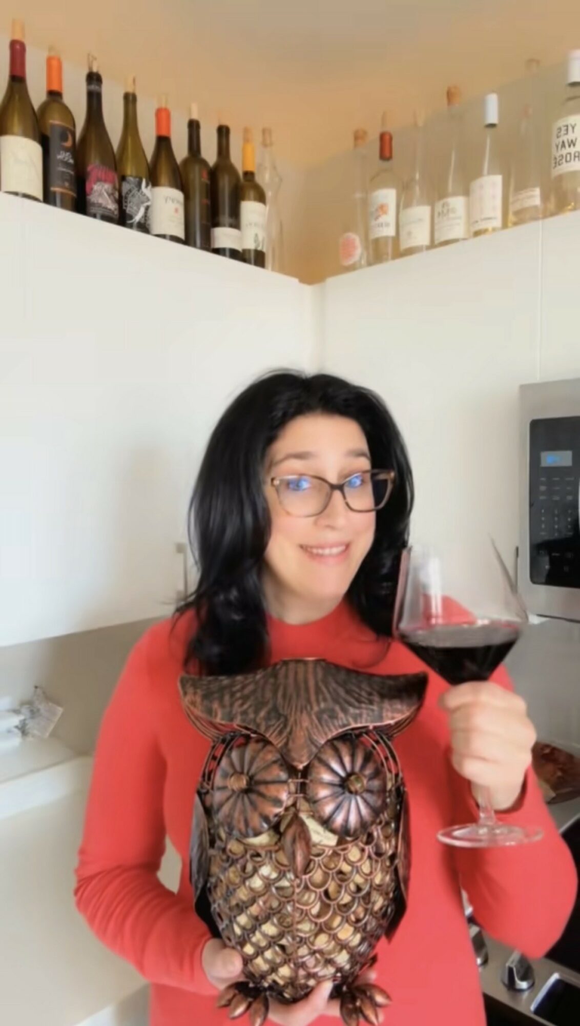 Woman with glasses holding a glass of red wine and an owl-shaped object, standing in front of a white wall with bottles on top.