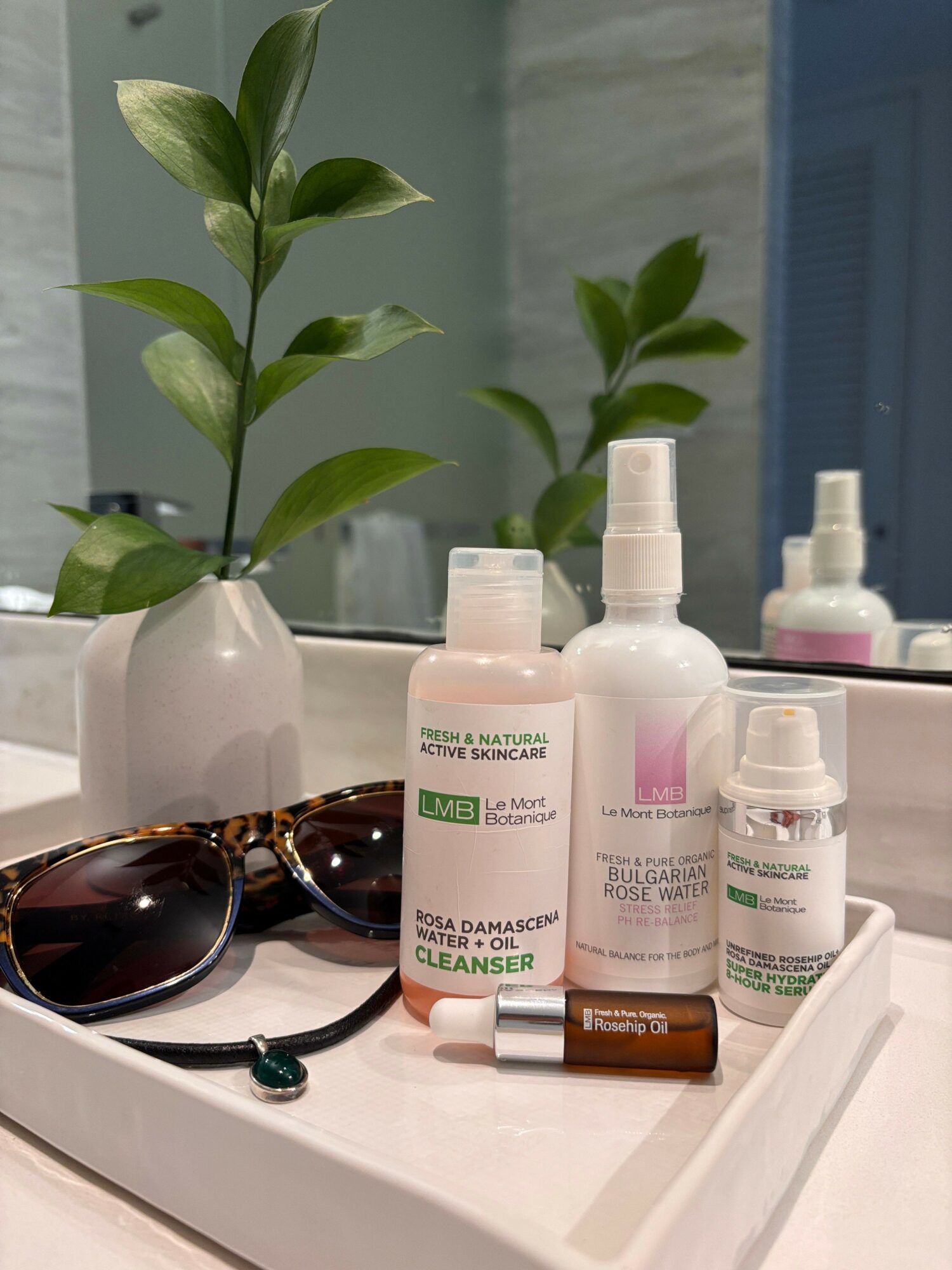 Various skincare bottles, a pair of sunglasses, and a small plant on a white surface, with a mirror in the background.