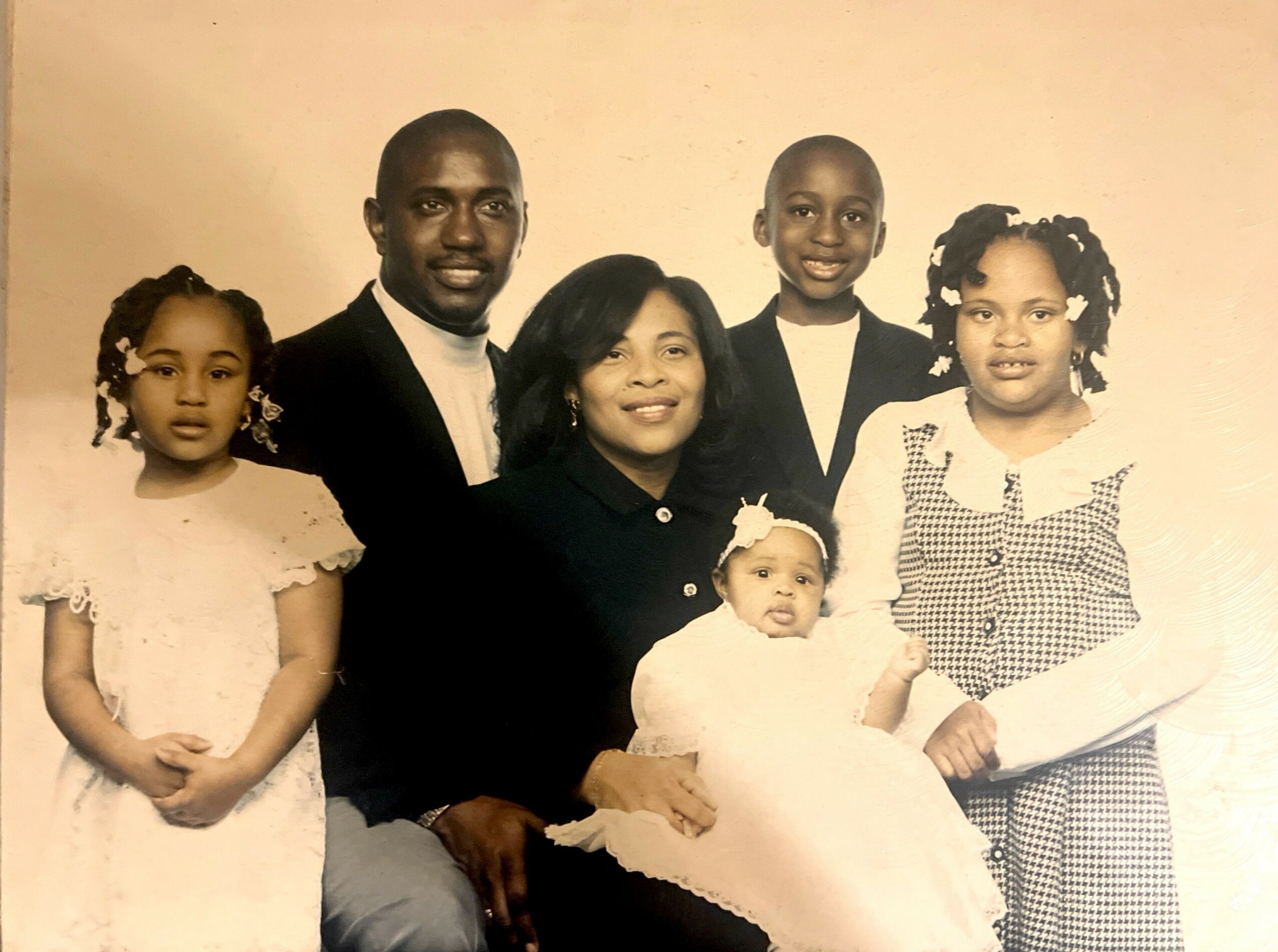 Family portrait with two adults and three children, all smiling, in a beige background, with the youngest baby in front.