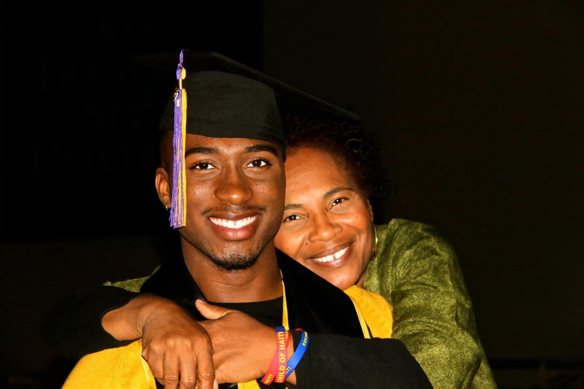 A young man in a graduation cap and gown smiling with a woman, both hugging, in a dark background.