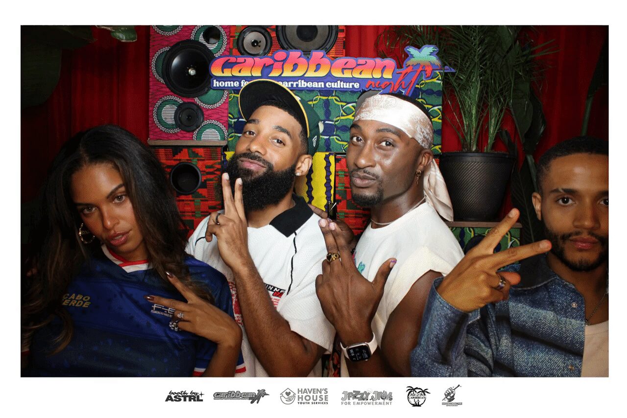 Four people posing together in front of a Caribbean-themed backdrop with speakers and plants, making hand gestures and facial expressions.