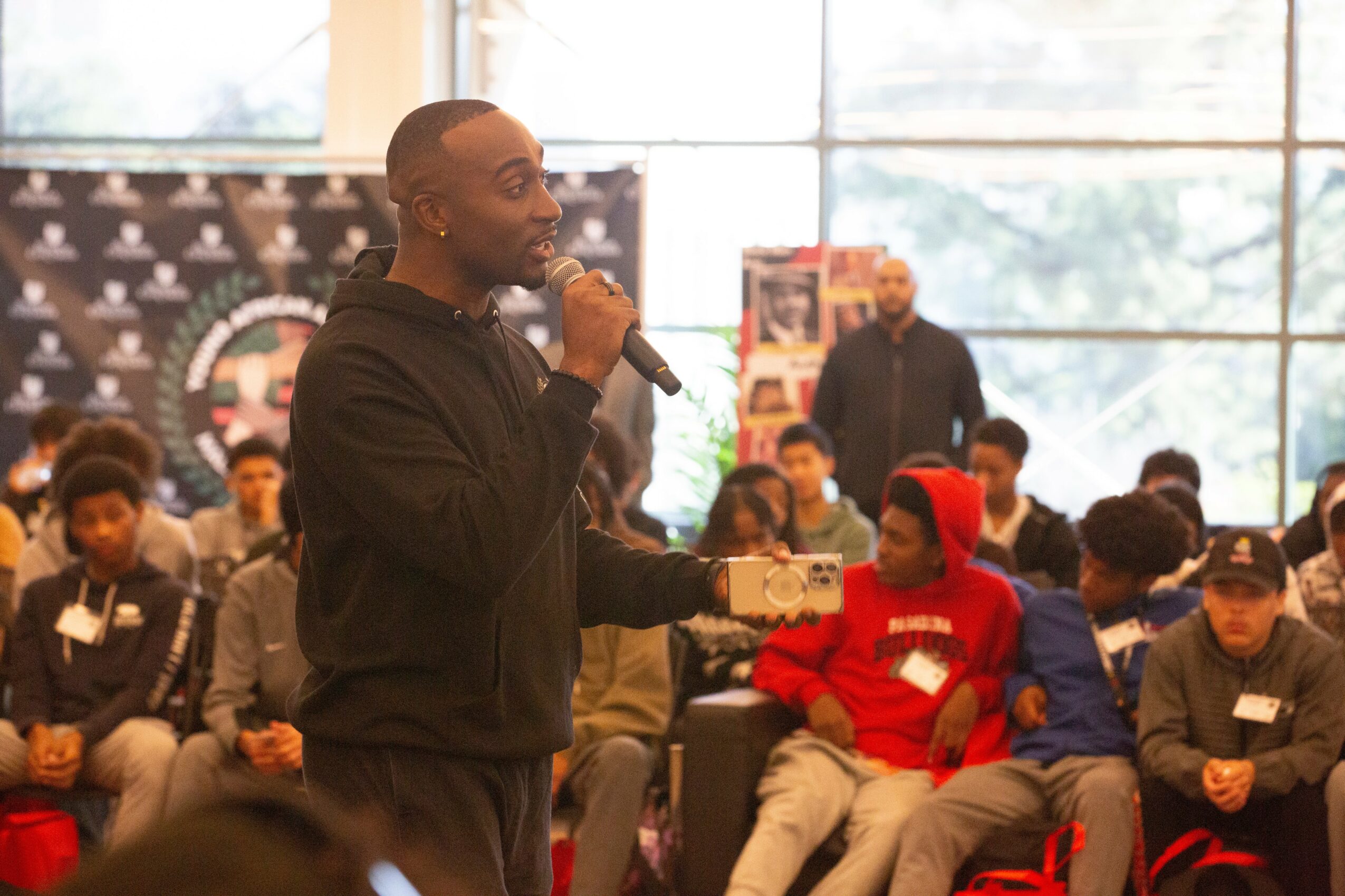 Man speaking into microphone in a large indoor gathering with seated audience members, some wearing hoodies and hats.