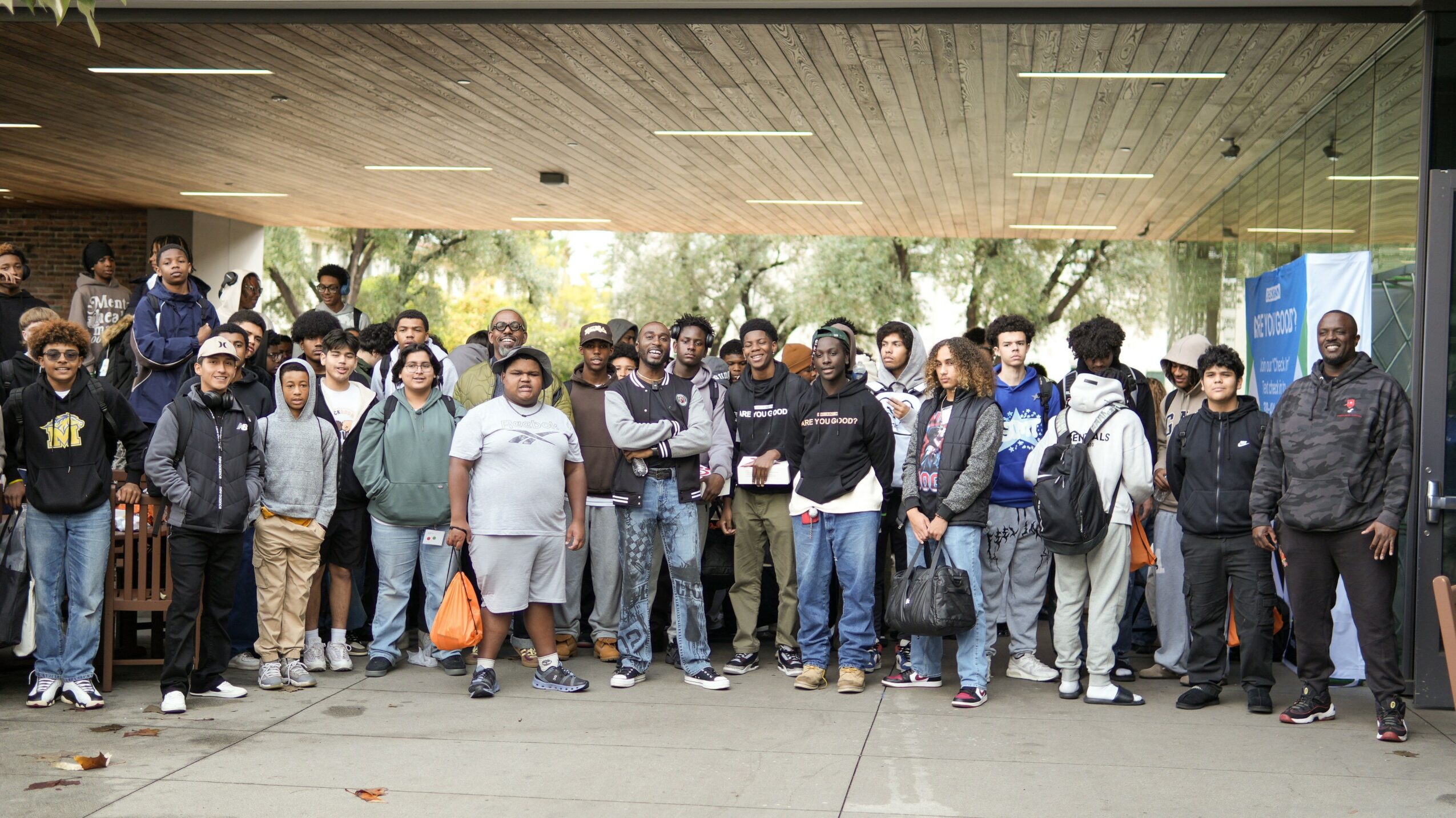 Group of people standing outdoors under a covered area, some holding bags and backpacks, with trees in the background.