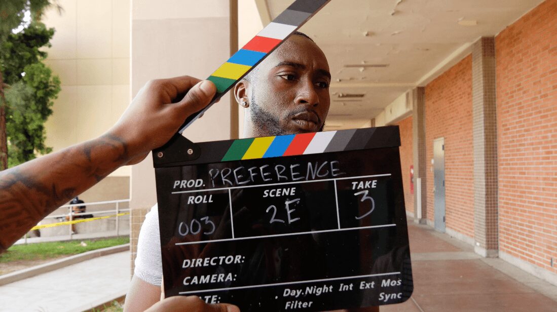 Man with a serious expression is filmed outdoors, holding a clapperboard, with a brick building and trees in the background.