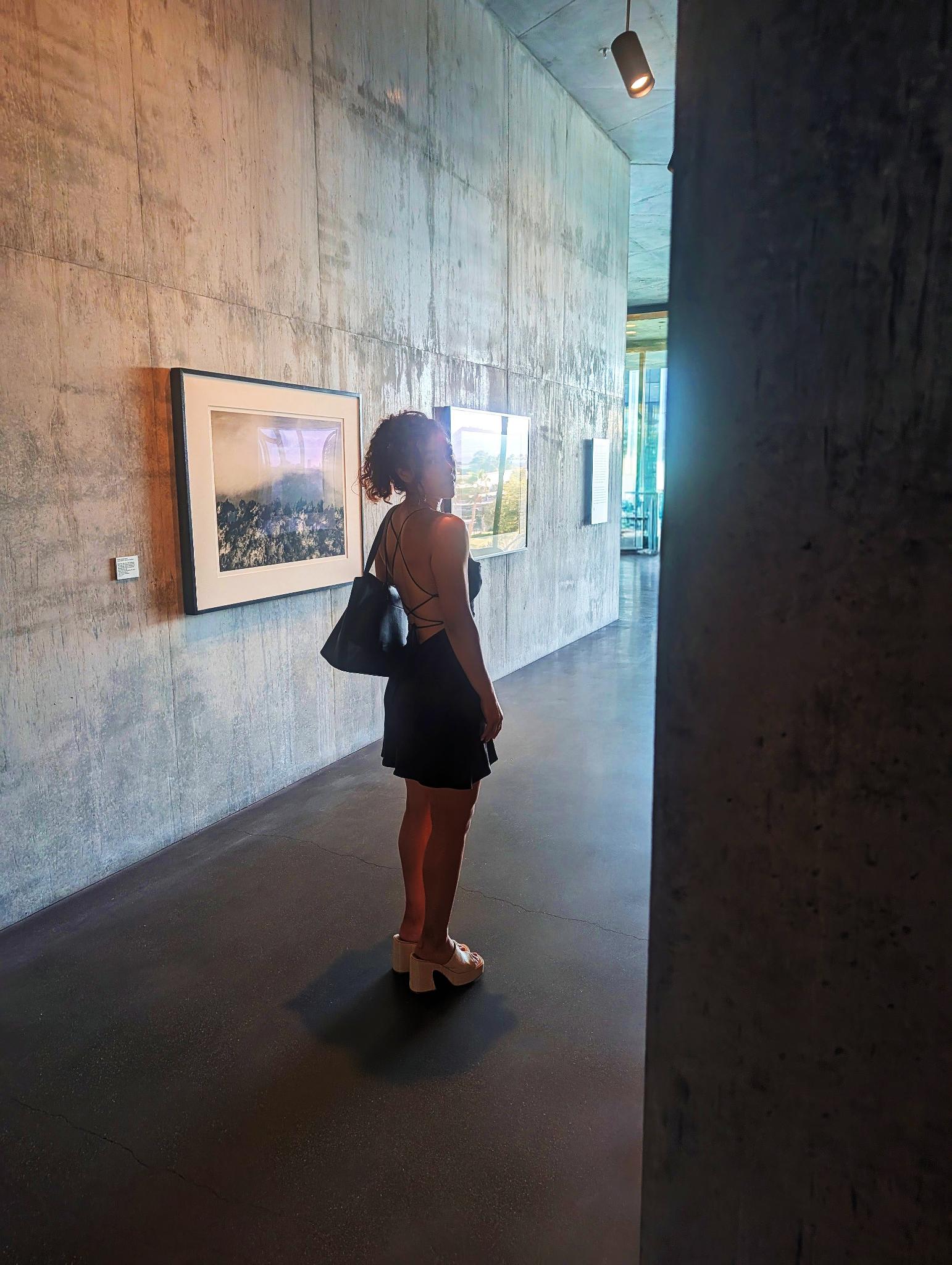 Person standing on a skateboard in an art gallery, looking at framed artwork on a concrete wall.