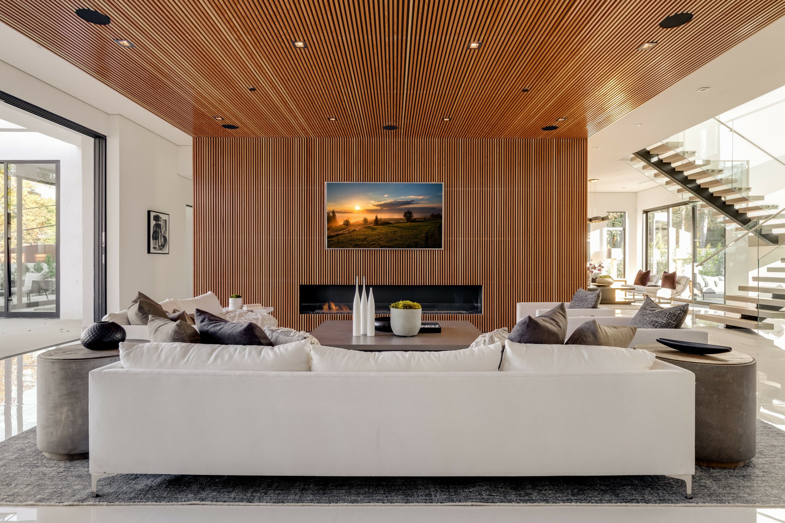 Modern living room with white sofa, wooden wall, TV, and large windows, featuring a staircase and natural light.