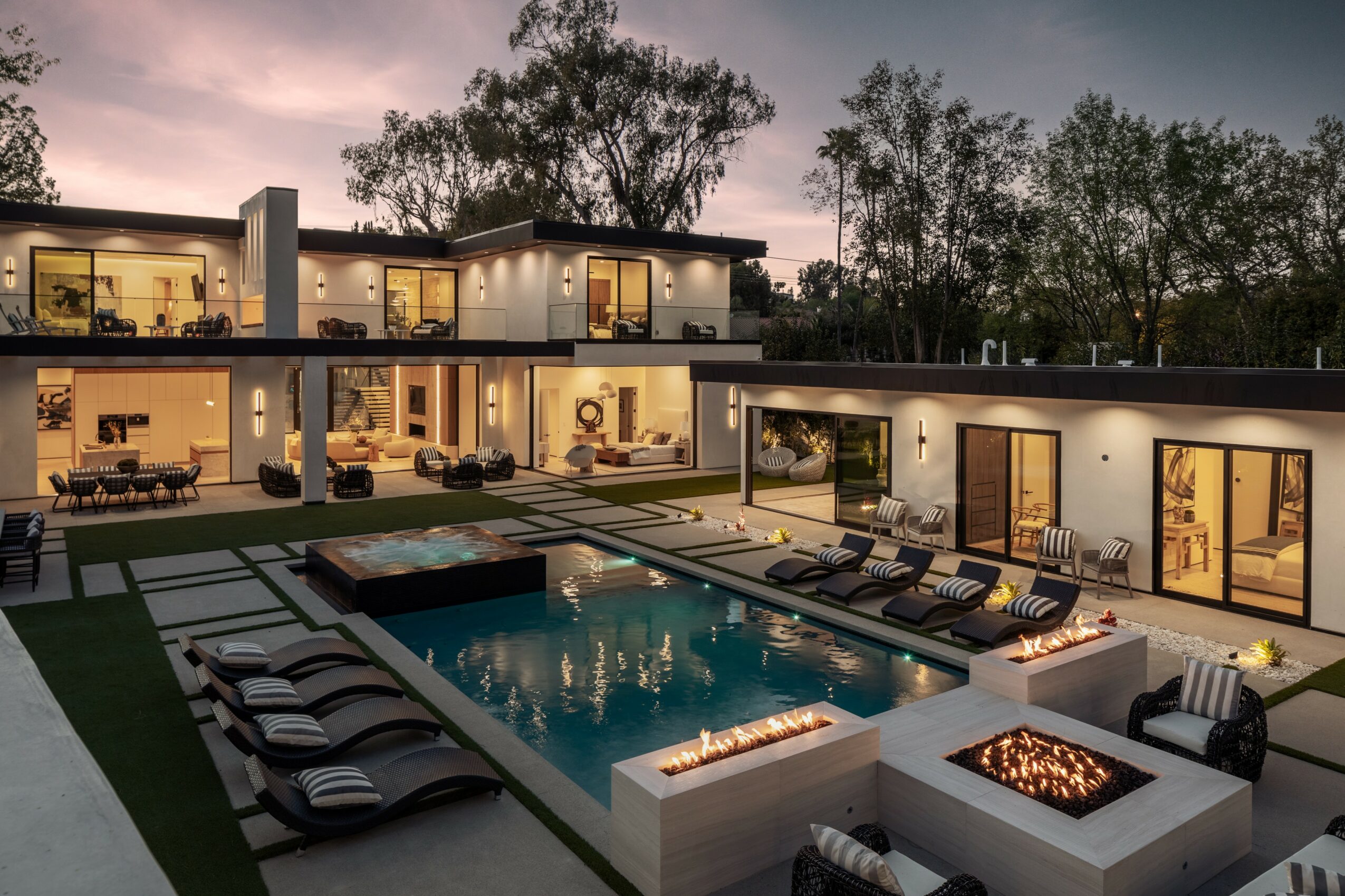 Modern two-story house with large windows, outdoor pool, lounge chairs, and fire pit, surrounded by trees at dusk.