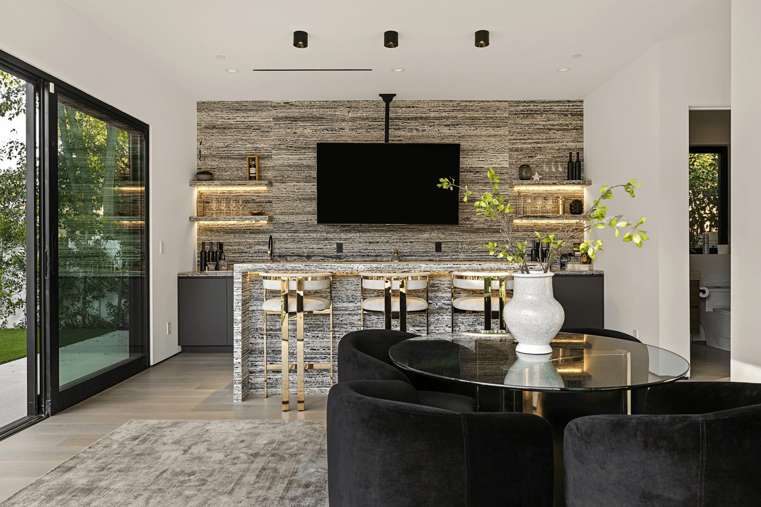 Living room with a stone accent wall, flat-screen TV, bar area, black round table with a white vase, and sliding glass door.