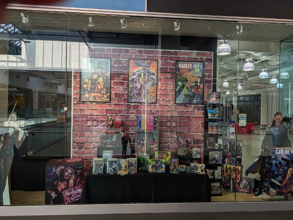 Store window display with comic books and posters against a brick wall, inside a shopping mall, with reflections on glass.