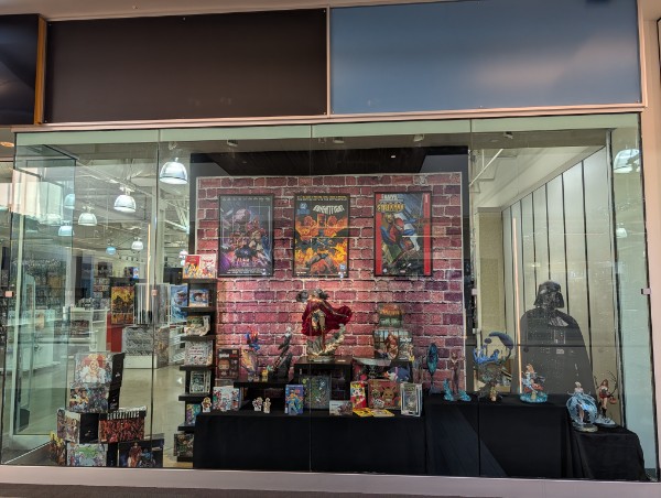 Store display with colorful posters, toys, and decorations against a brick wall background, viewed through glass windows.