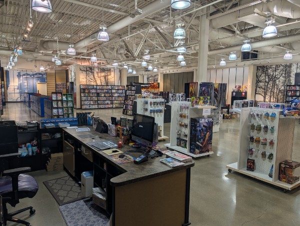 View of a retail store interior with shelves, checkout counter, and overhead lighting, organized with various products displayed.