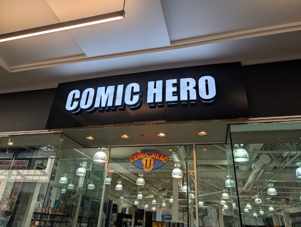 Storefront sign reads 'COMIC HERO' in large illuminated letters, with a glass entrance below and store interior visible.