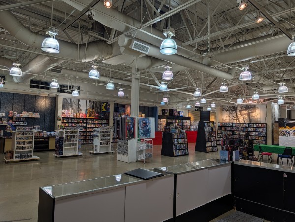 Interior of a retail store with shelves, displays, and overhead lighting fixtures, viewed from a counter area.