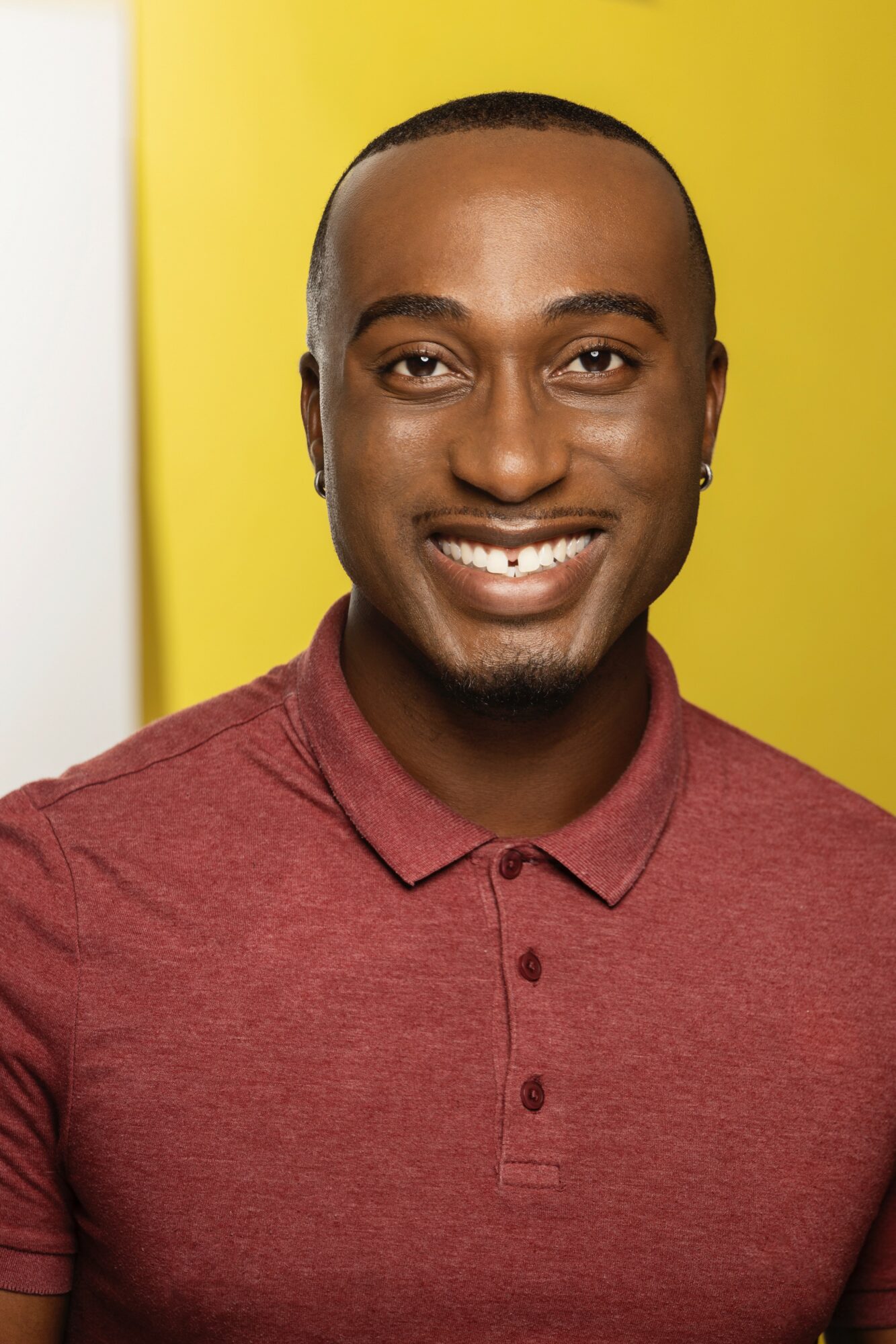 Portrait of a smiling man with a yellow background, wearing a maroon polo shirt.