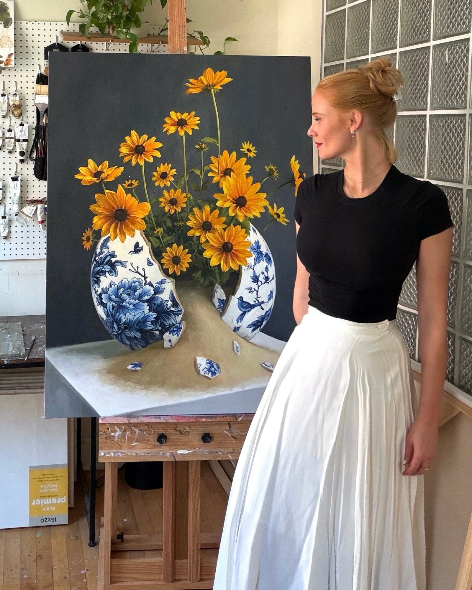 Red-haired woman in black top and white skirt looking at a still life painting of yellow flowers in blue and white vases.