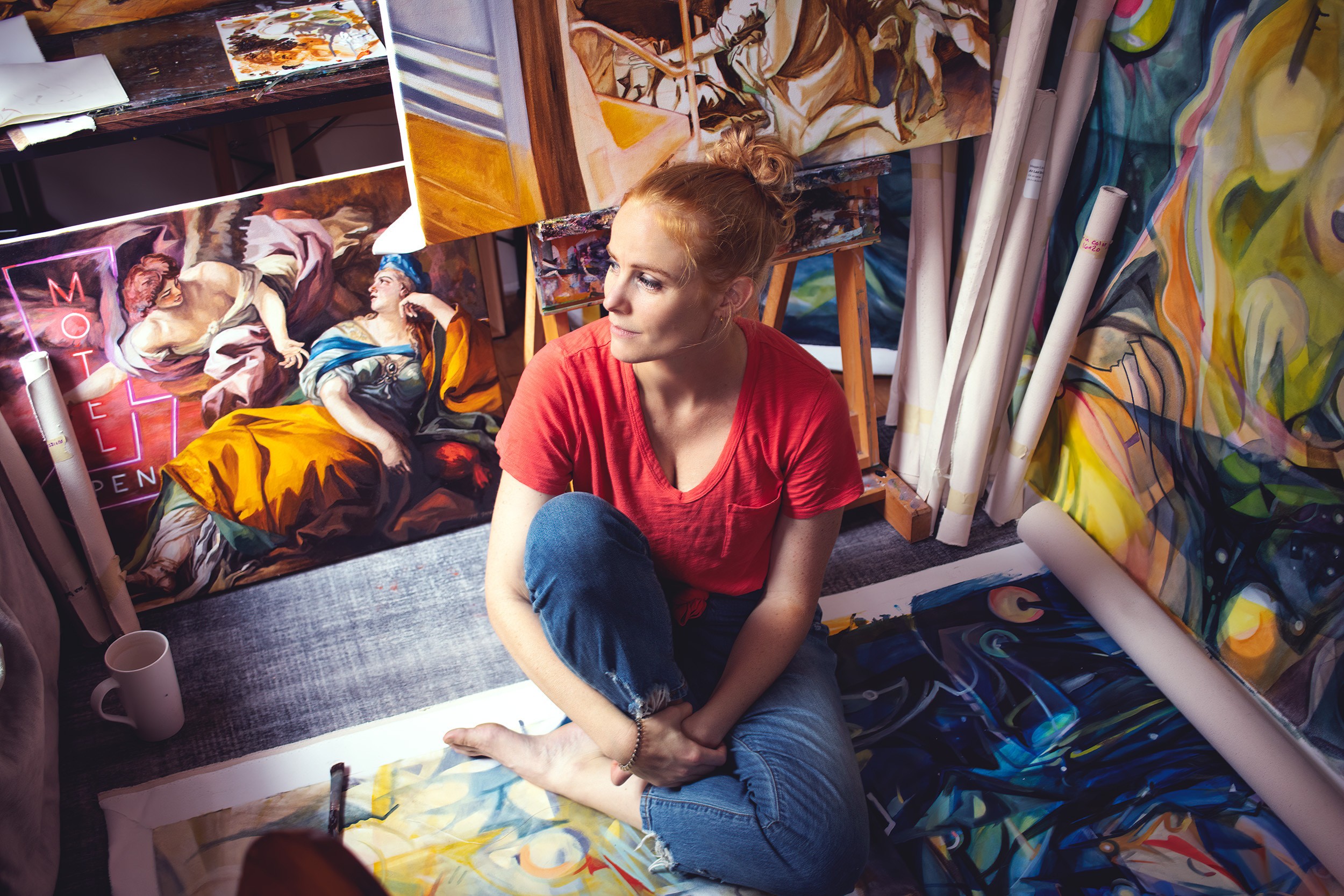 Young woman sitting on the floor surrounded by colorful paintings and art supplies in an art studio.