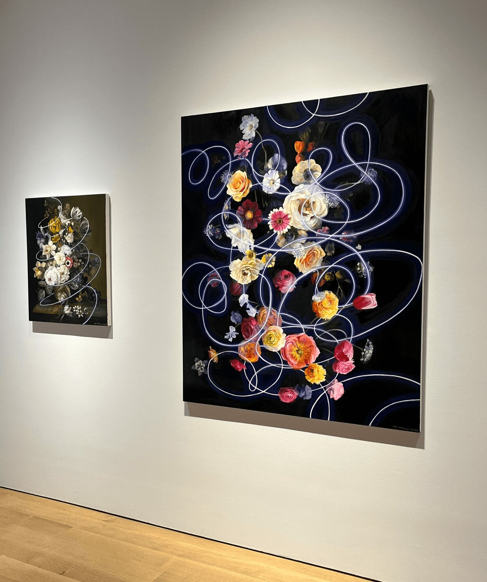 Two floral paintings on a white gallery wall, with the larger one featuring colorful flowers and swirling lines on a black background.