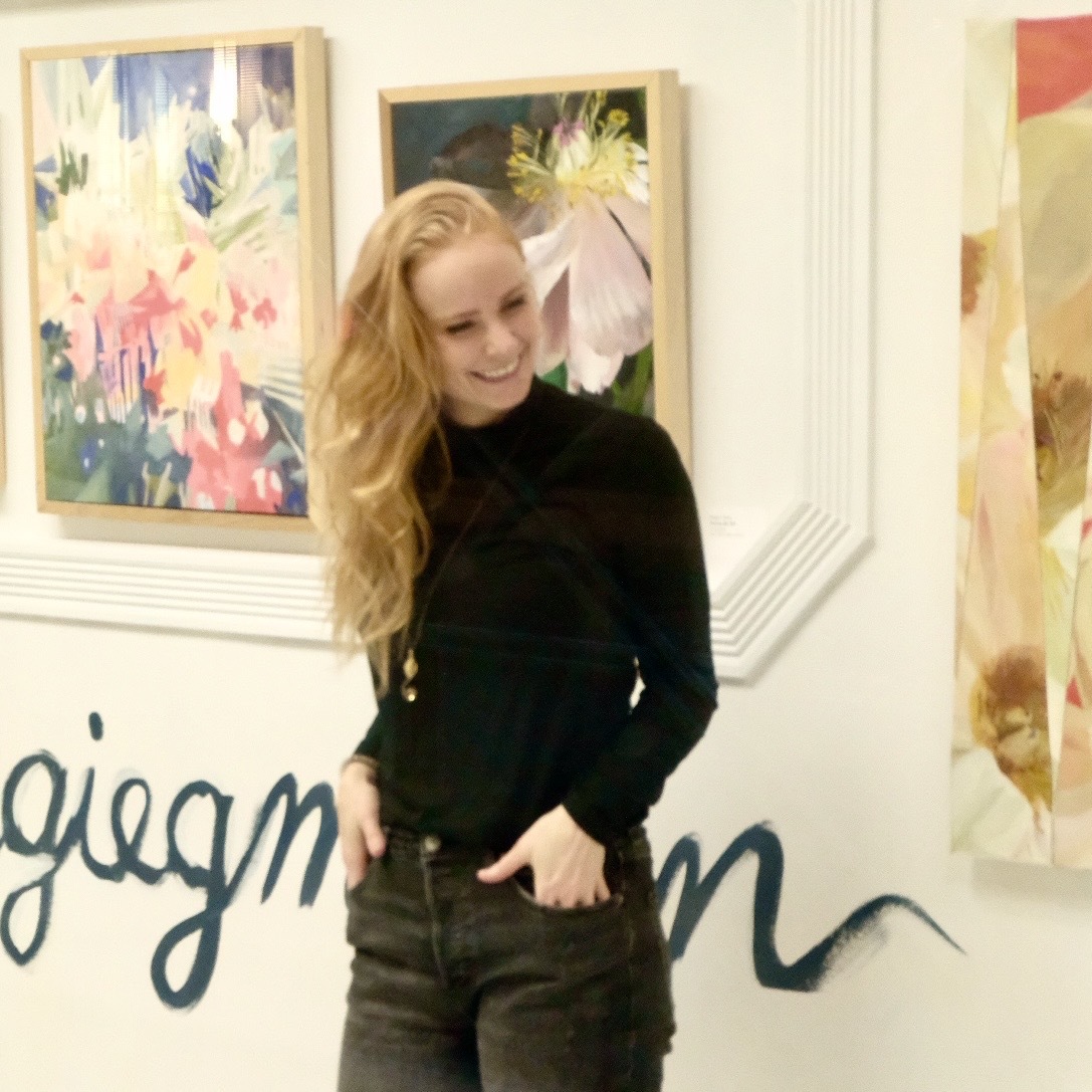 Young woman with long, wavy blonde hair smiling, wearing a black top and dark jeans, standing in an art gallery.