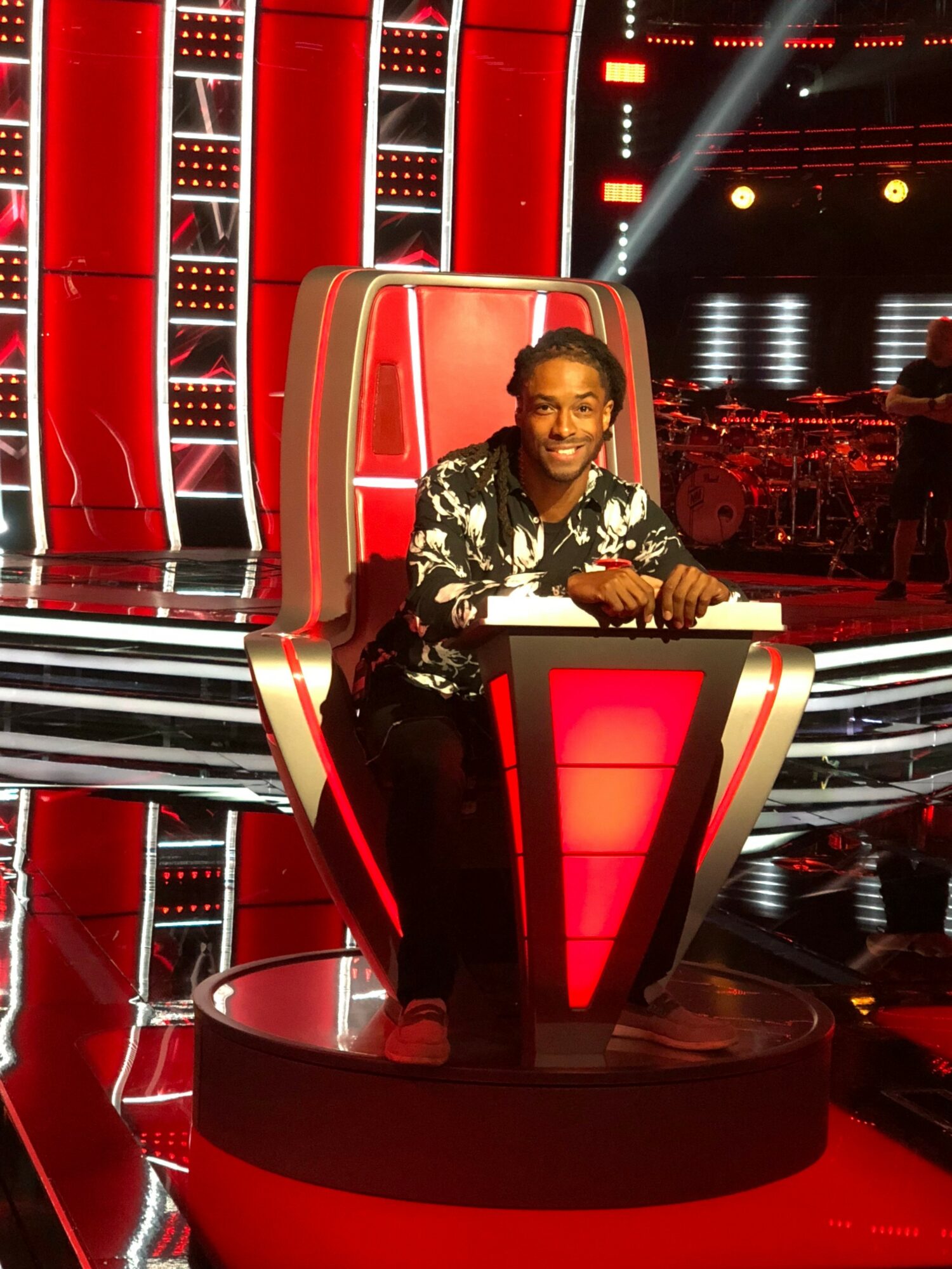 Person sitting in a red and black chair on a stage with bright lights and a large screen behind.