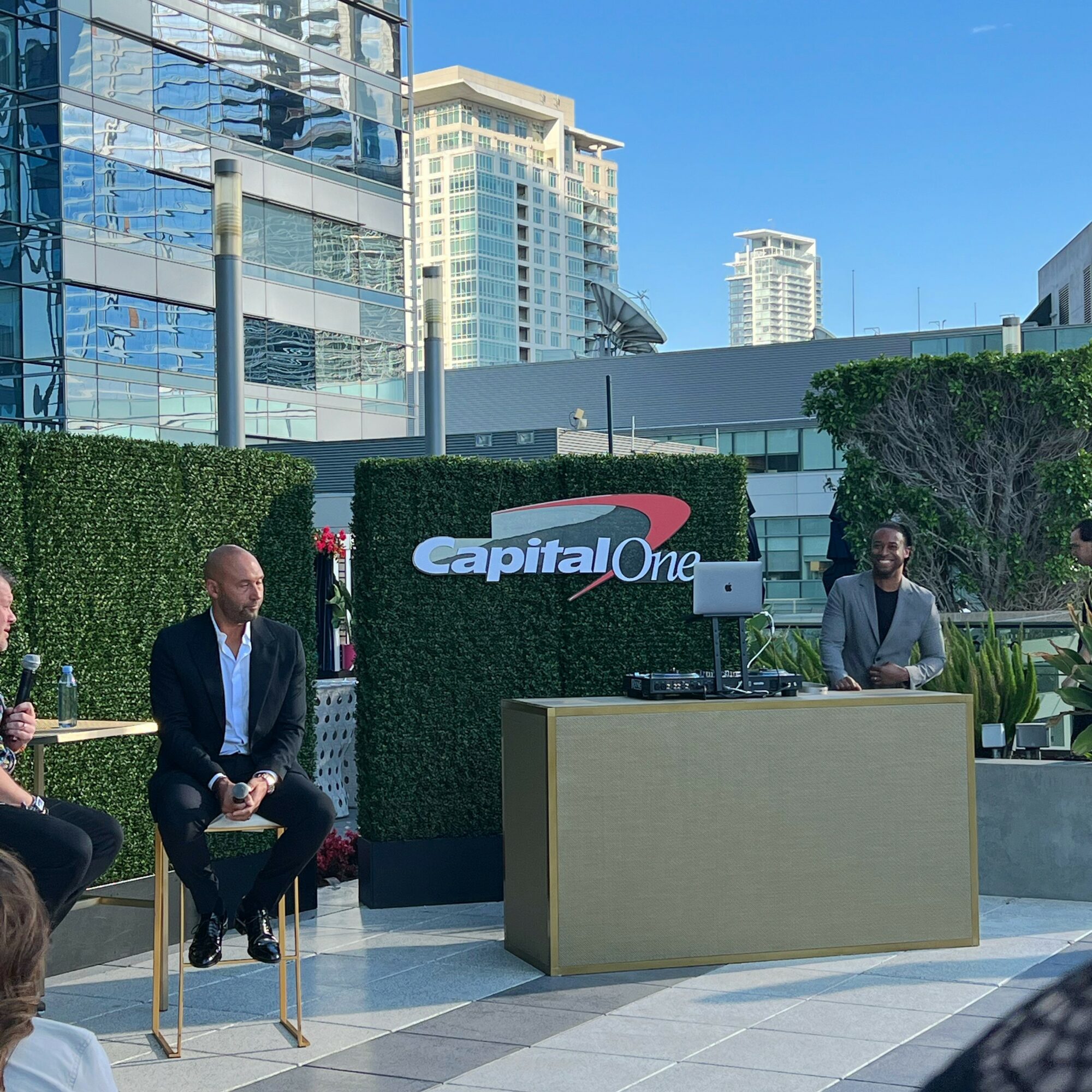 Two men in suits sit outdoors near a DJ setup with a Capital One sign, city buildings in background.